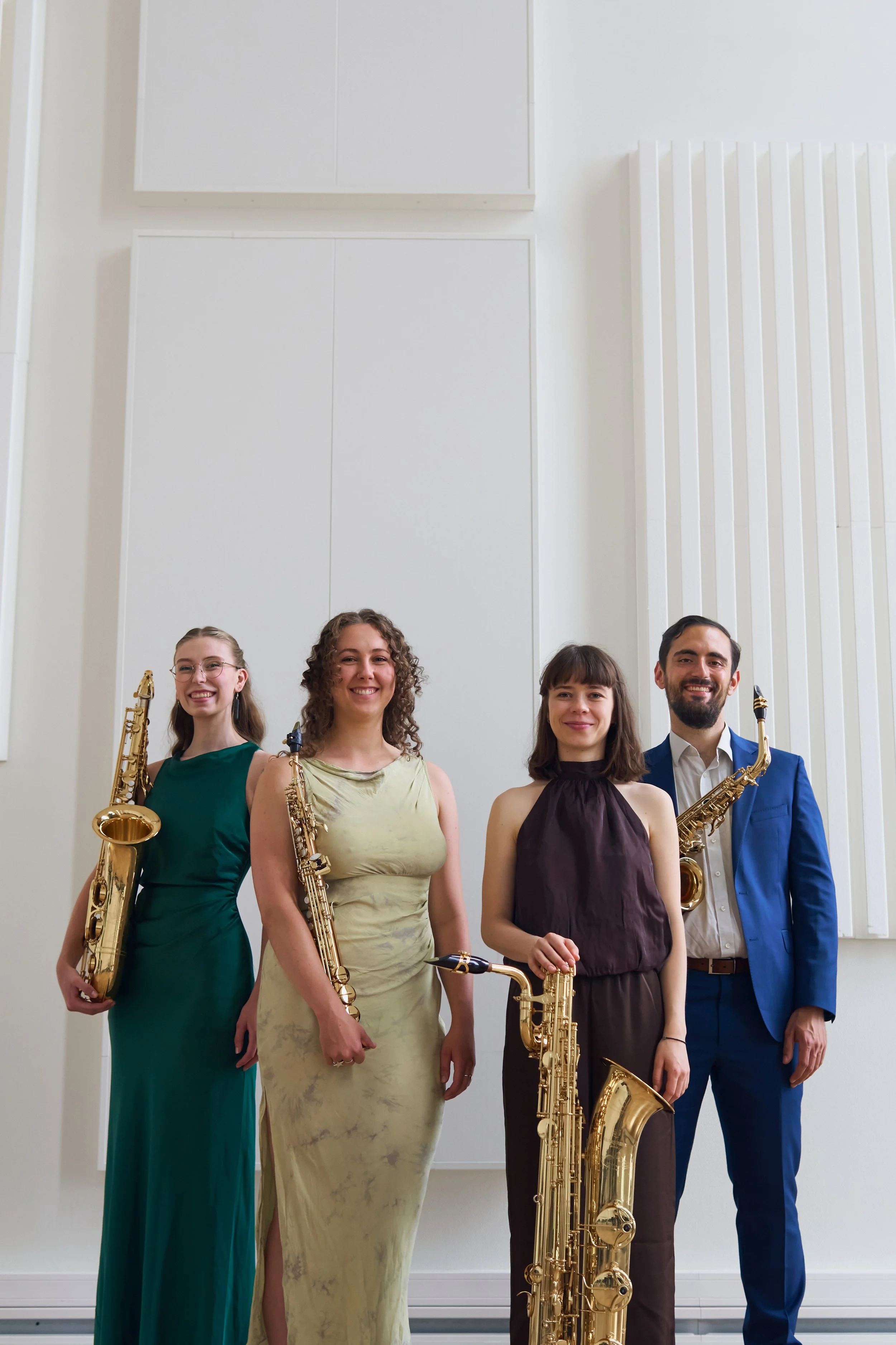 Group of five musicians standing in a row, holding saxophones, in a bright, modern room with white walls and high ceilings.