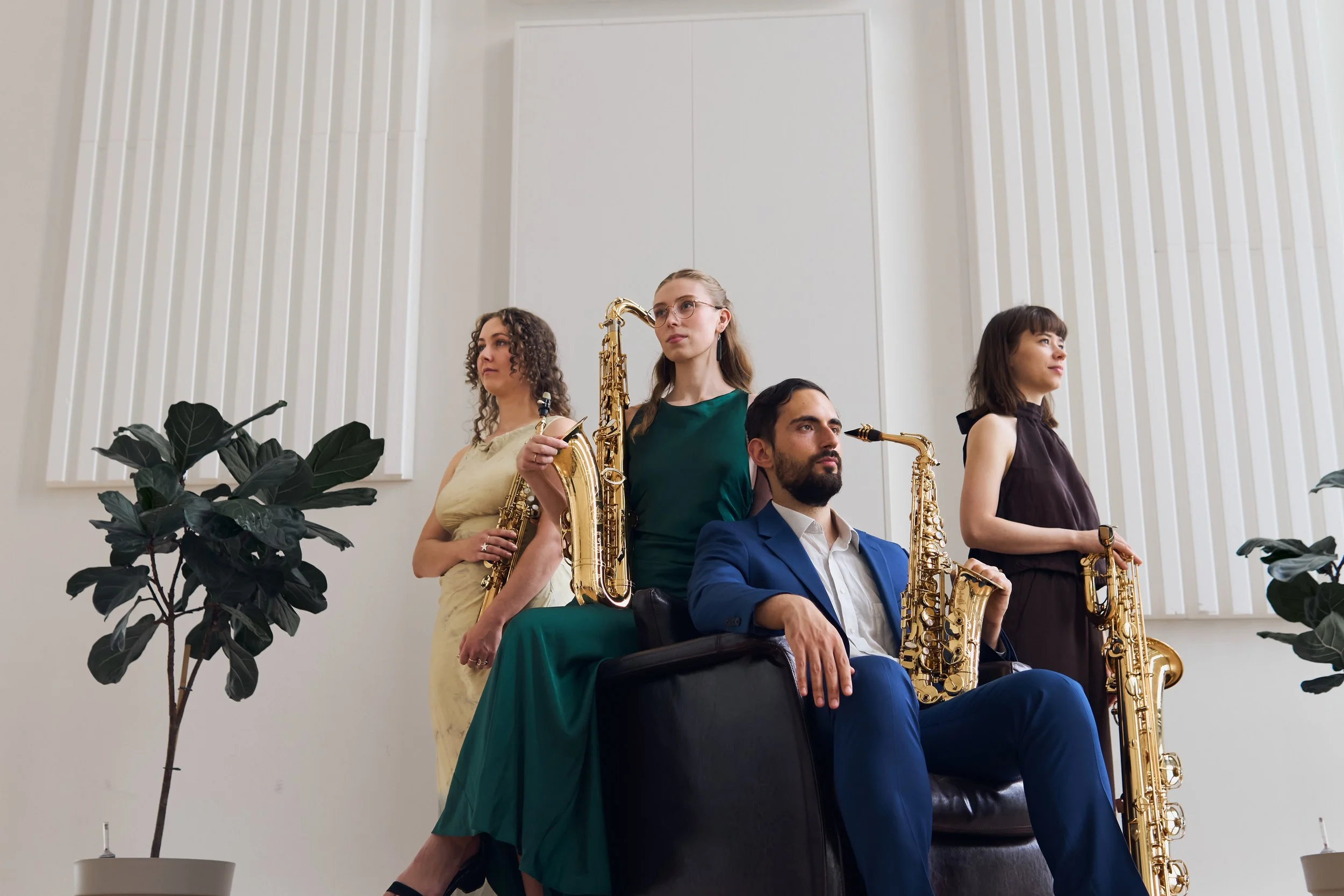 Four musicians, three women and one man, are holding or sitting near saxophones in an indoor setting against a white wall with vertical panels. Two large potted plants are on either side of the group.