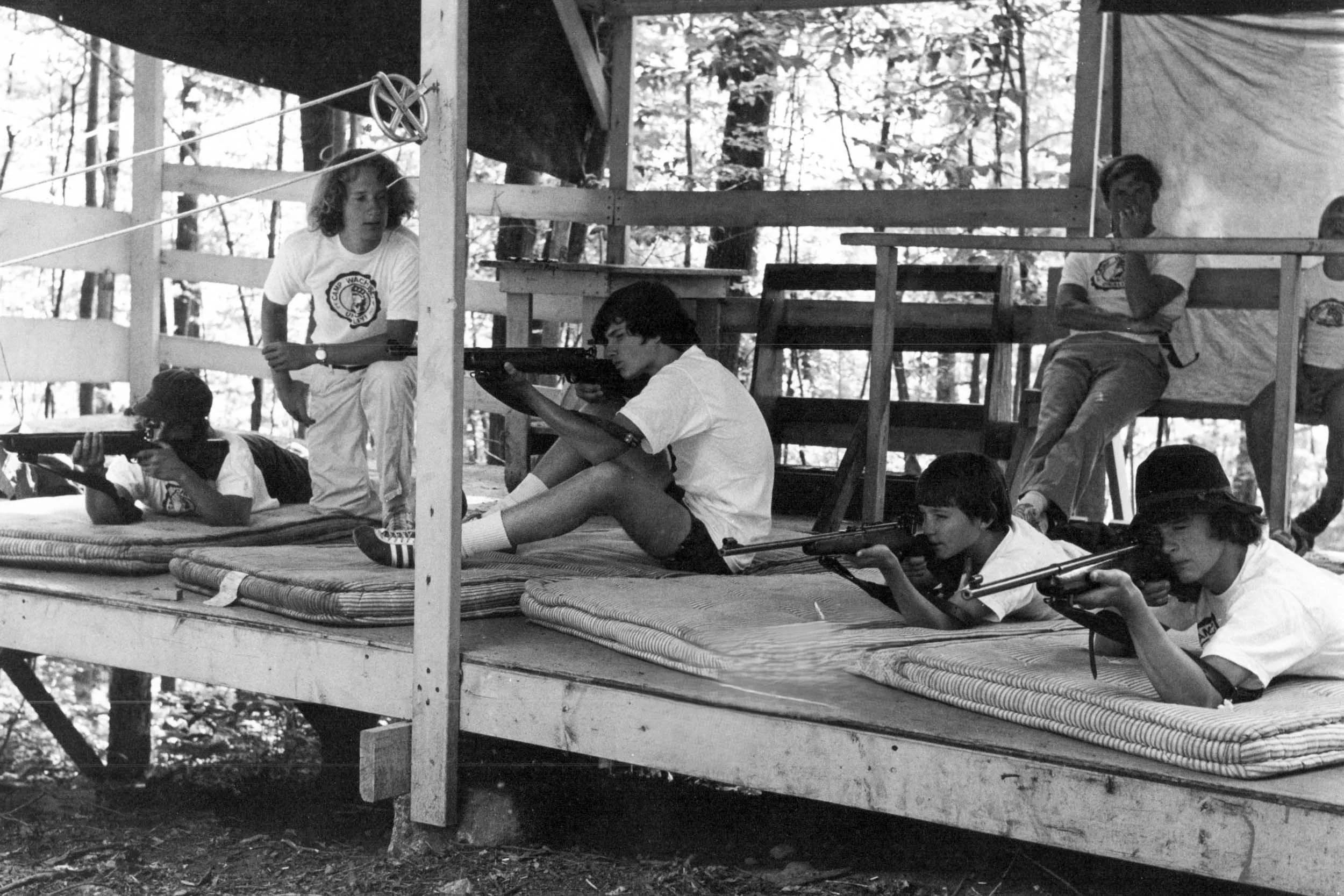 Black and white photo of boys at riflery range, aiming at targets.