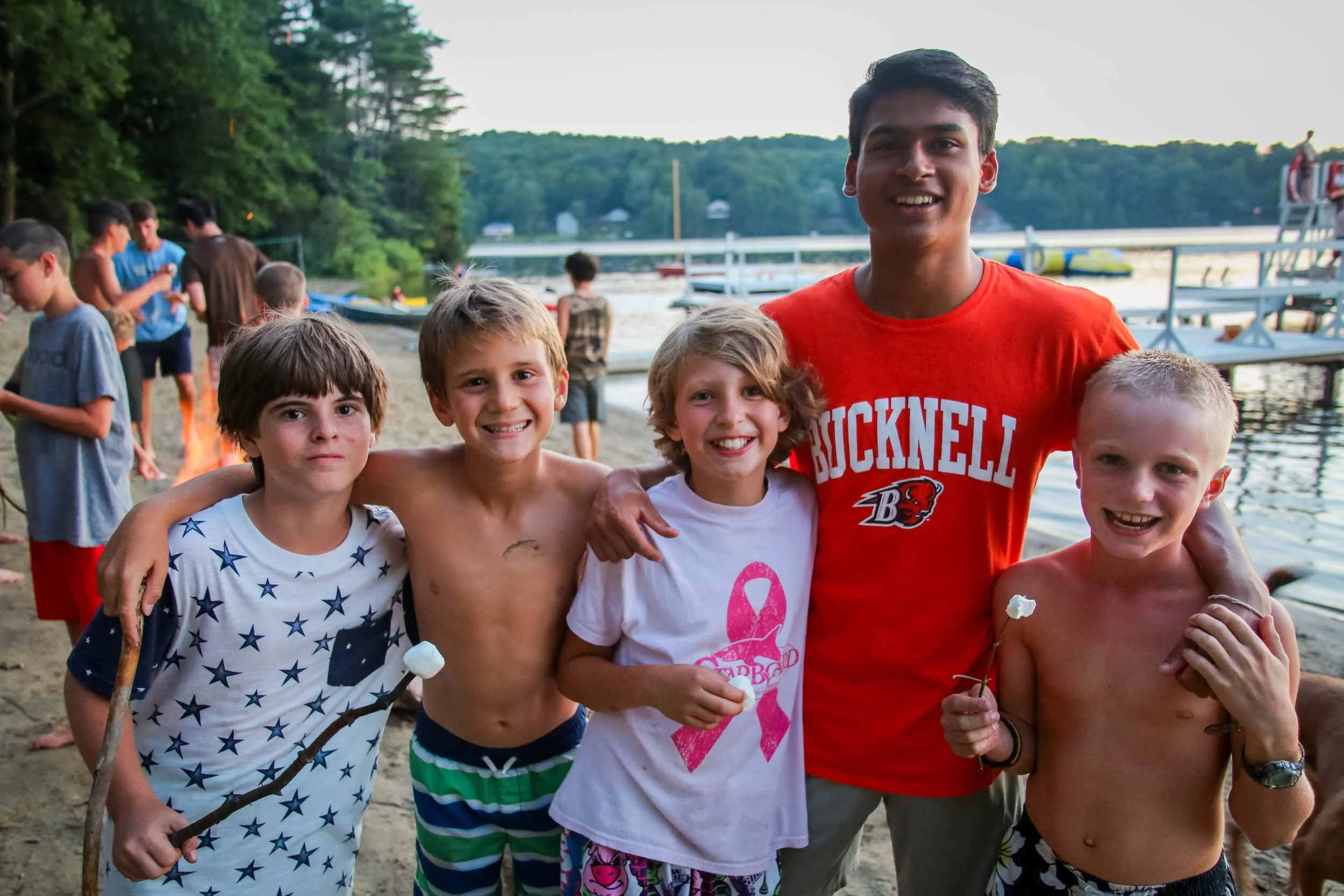 Four smiling boys and their counselor standing together on a beach holding sticks with marshmallows.