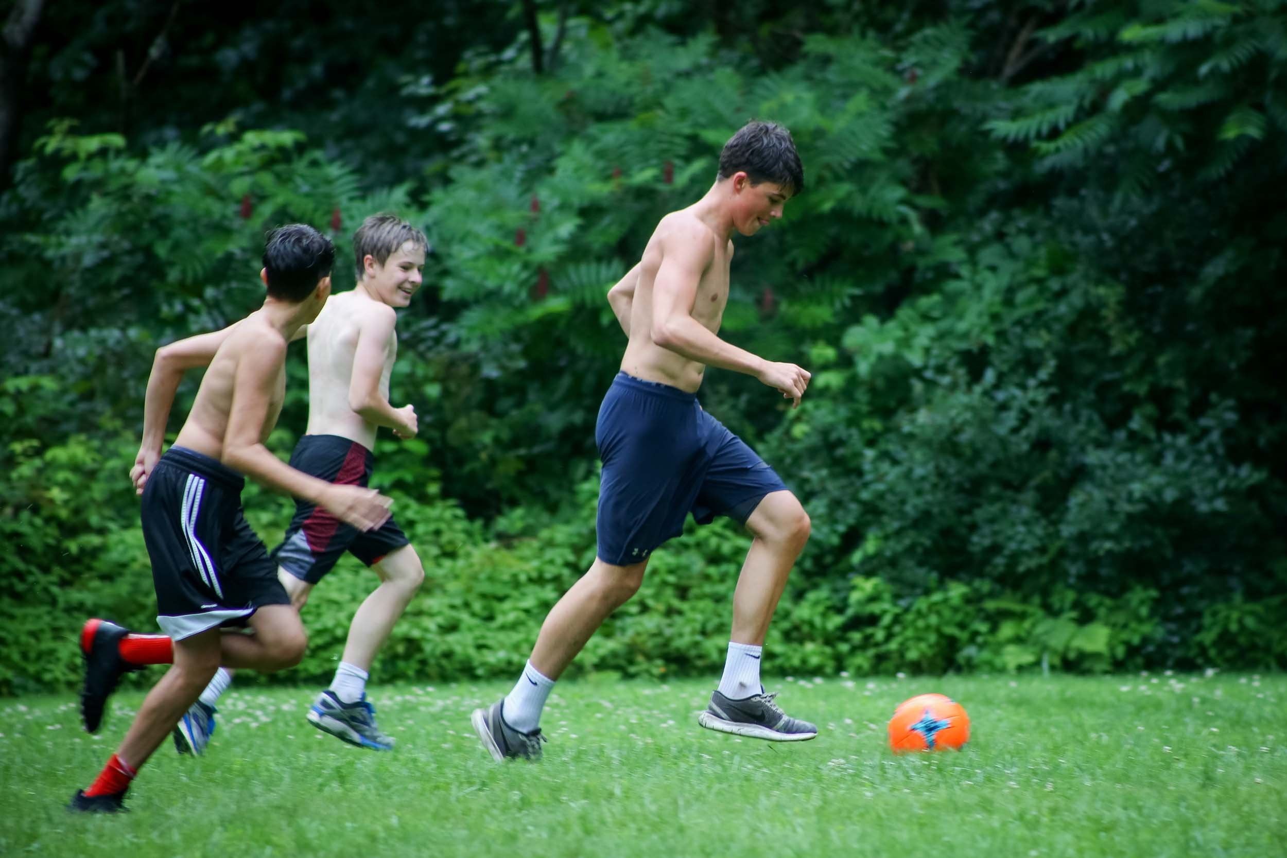 Three boys smiling play soccer outdoors near a forest.
