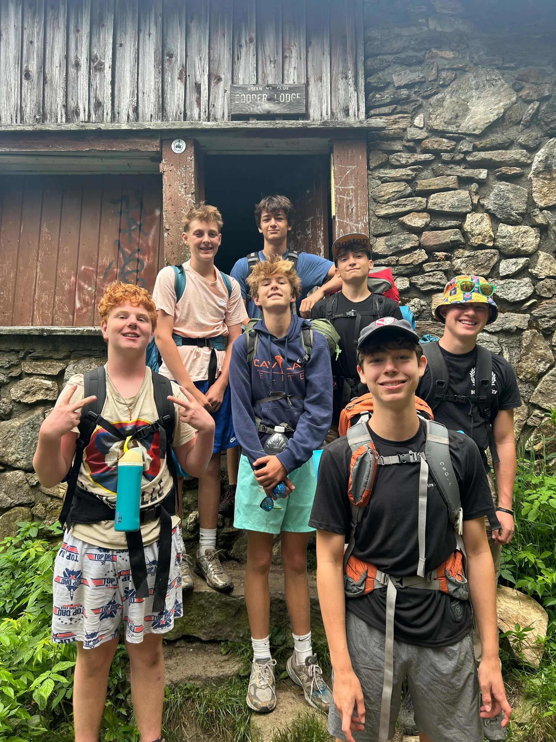 Group teen boys with backpacking gear standing in front of a rustic stone building during a hike.