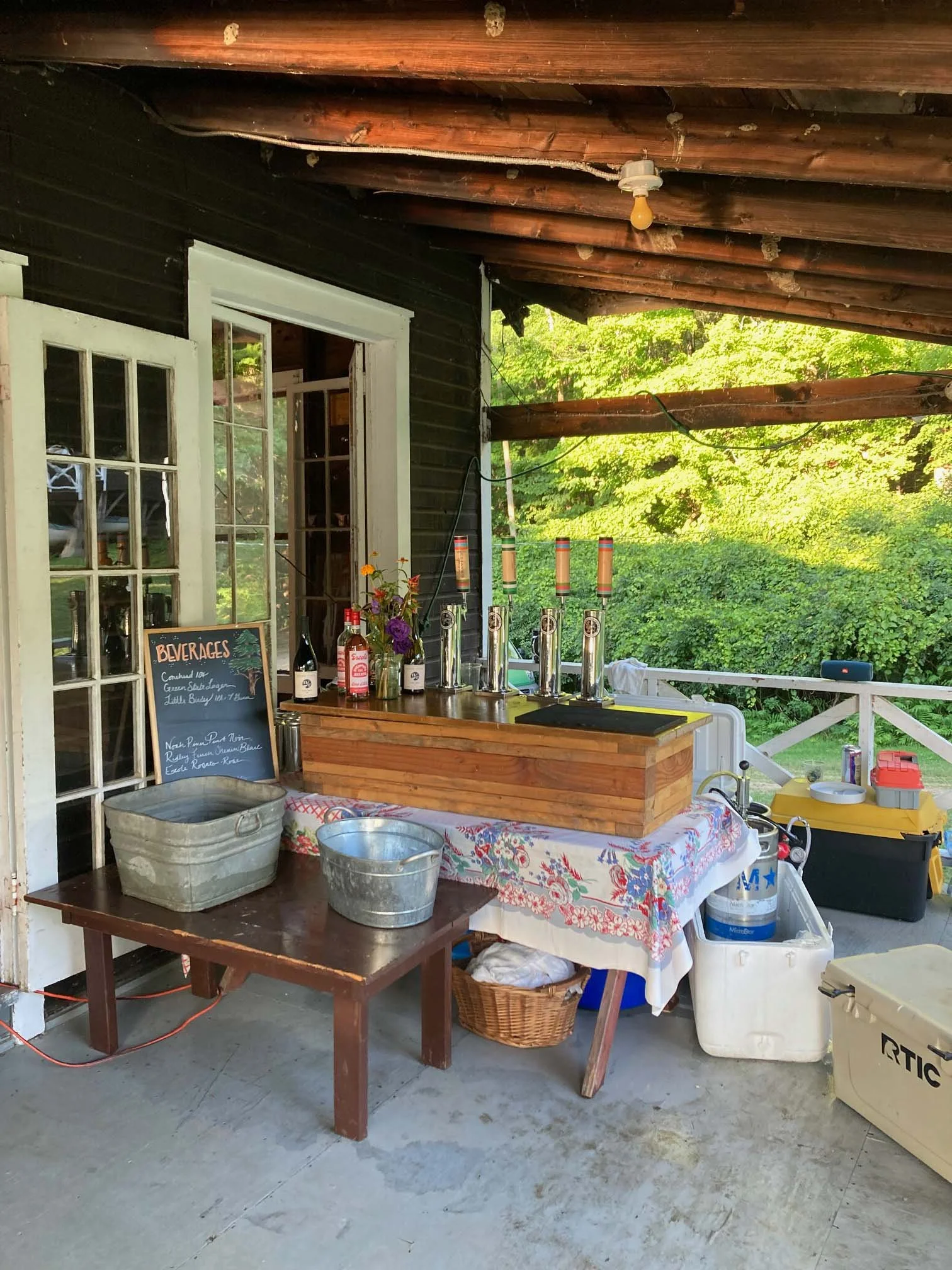 An outdoor bar setup with four beer taps on a wooden counter, a chalkboard menu, bottles, flowers.