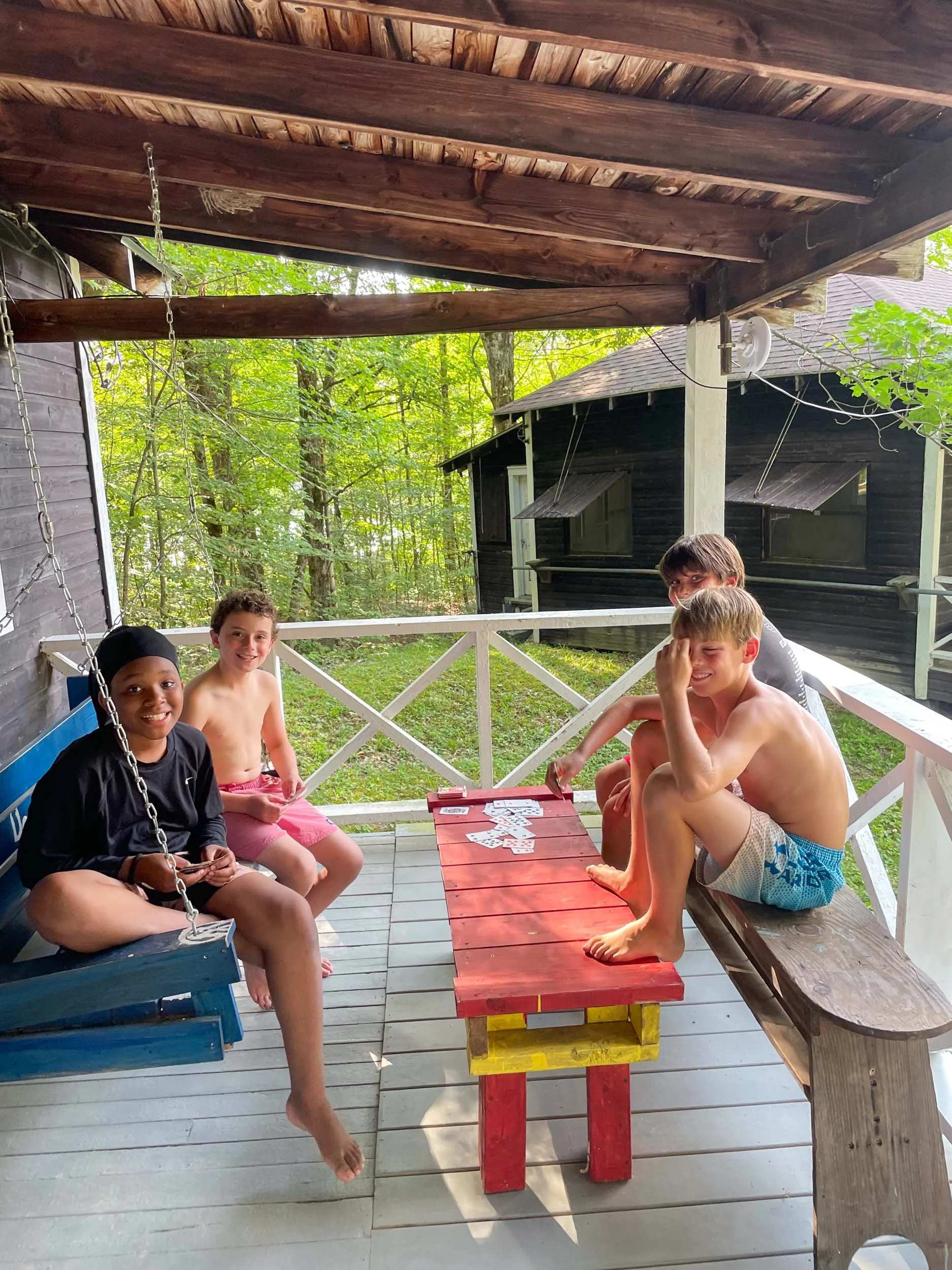 Four boys sitting on their cabin porch with a swinging bench, playing cards.