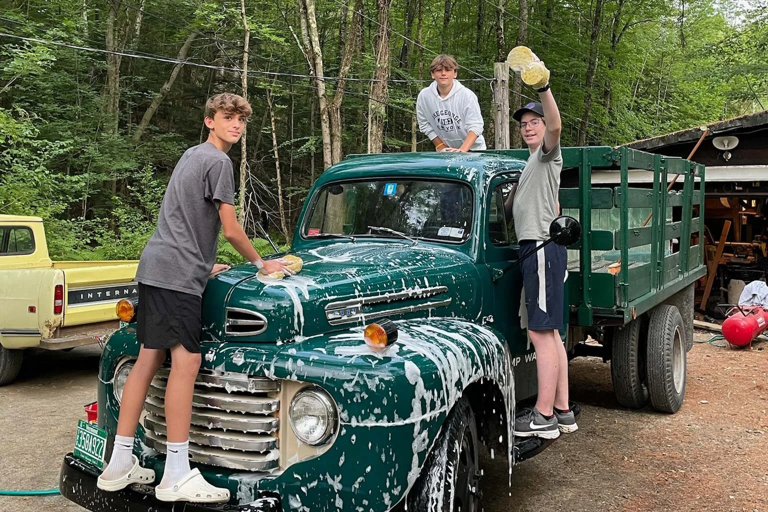 Three boys cleaning a green vintage truck with soap and water outdoors.