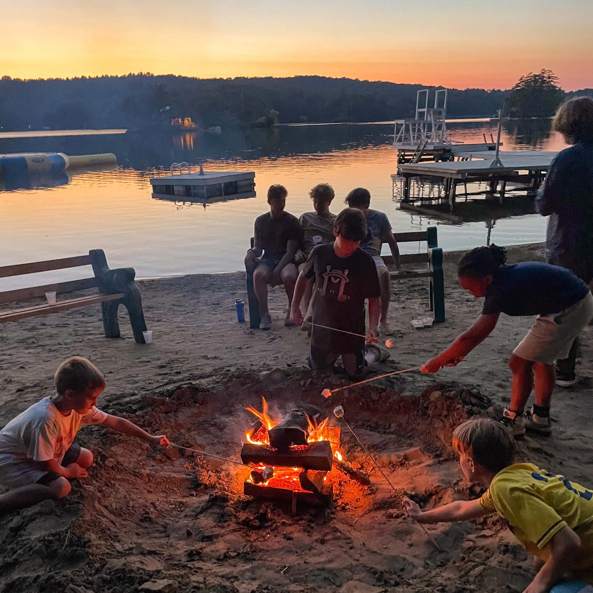 Campers sitting around a campfire on a sandy lakeside beach during sunset.