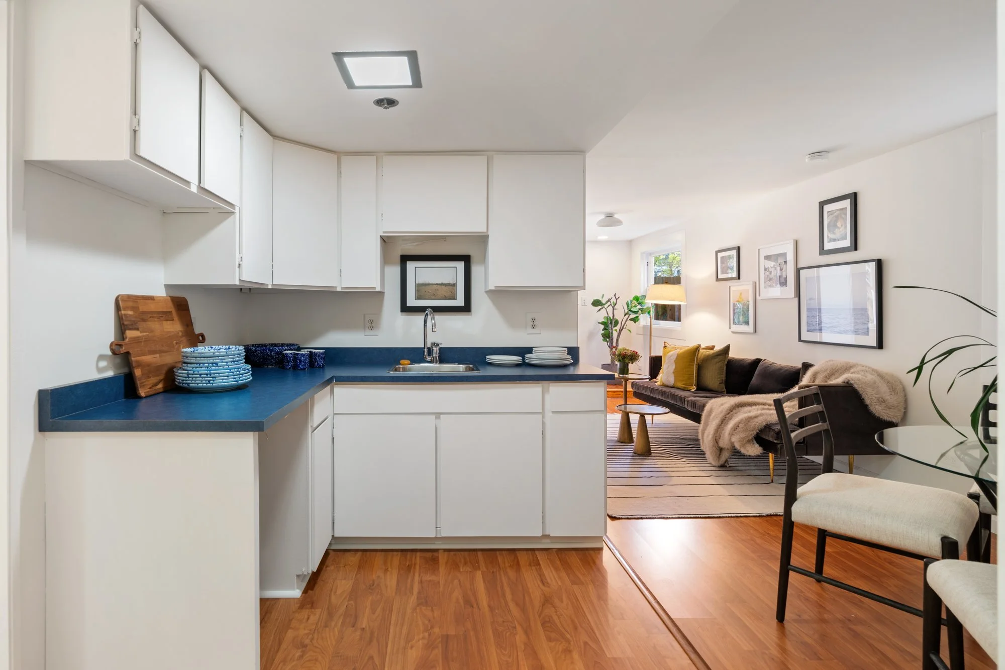 Open-concept living room and kitchen with hardwood floors, white cabinets, blue countertops, framed wall art, and seating area with a table, pillows, and throws.
