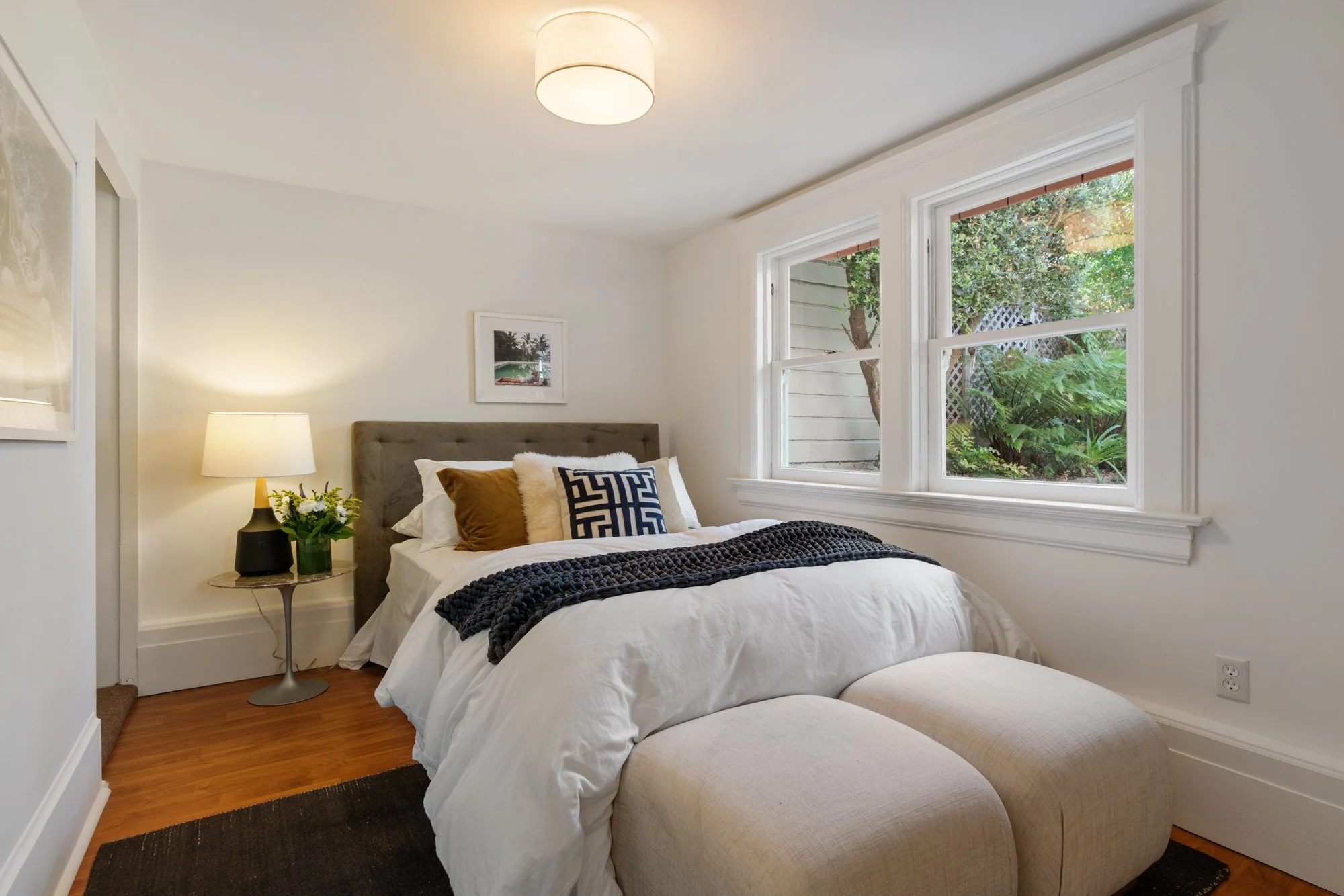 A cozy bedroom with a bed featuring white linens, decorative pillows, and a textured black throw. A beige ottoman is at the foot of the bed. To the left, a round side table with a black lamp and a vase of flowers. The room has white walls, a ceiling-