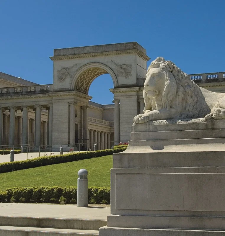 Statue of a reclining lion in front of a classical building with arch and columns.