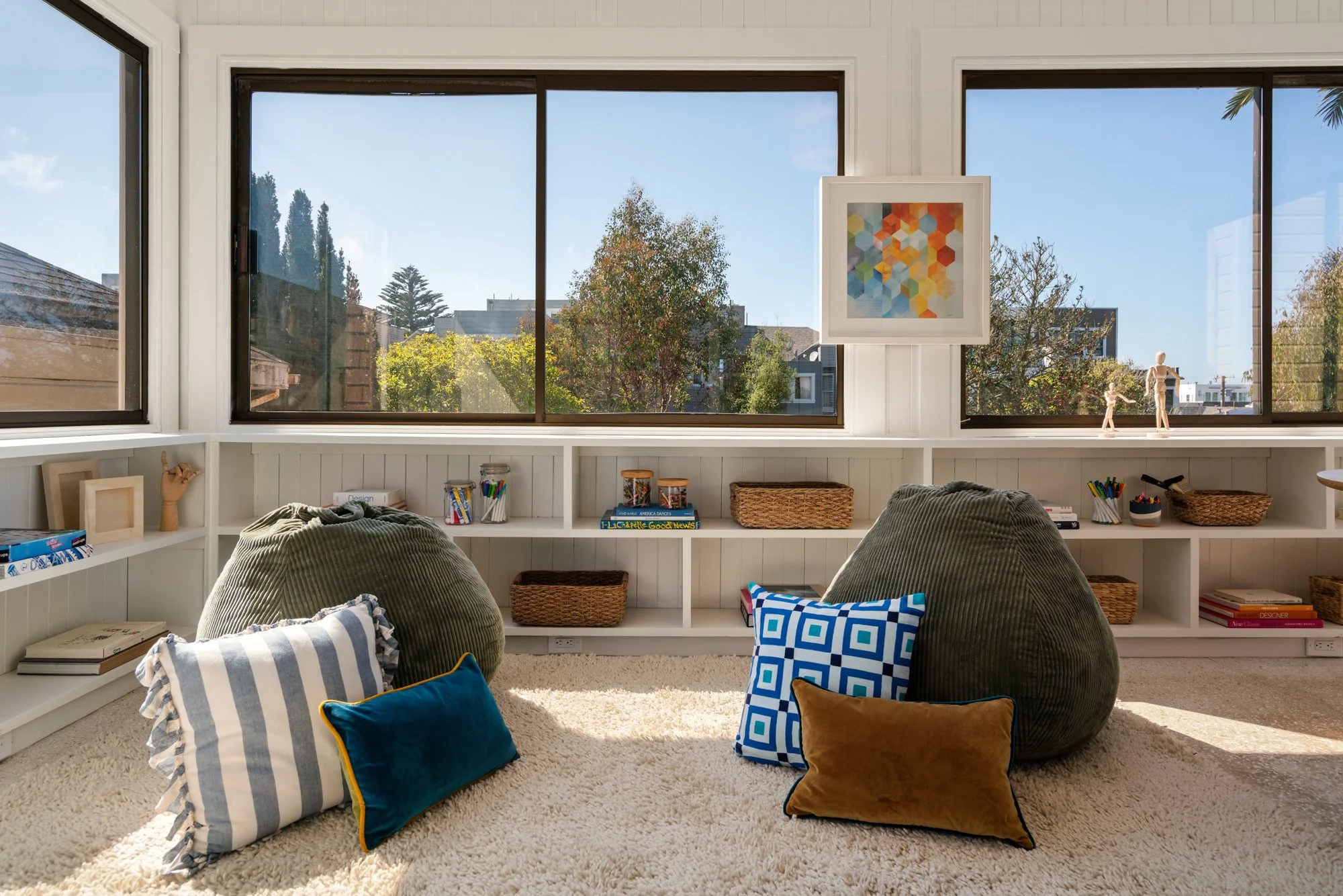 Sunlit living room with large windows, white built-in shelves filled with books and decor, and plush chairs with cushions on cream carpet.