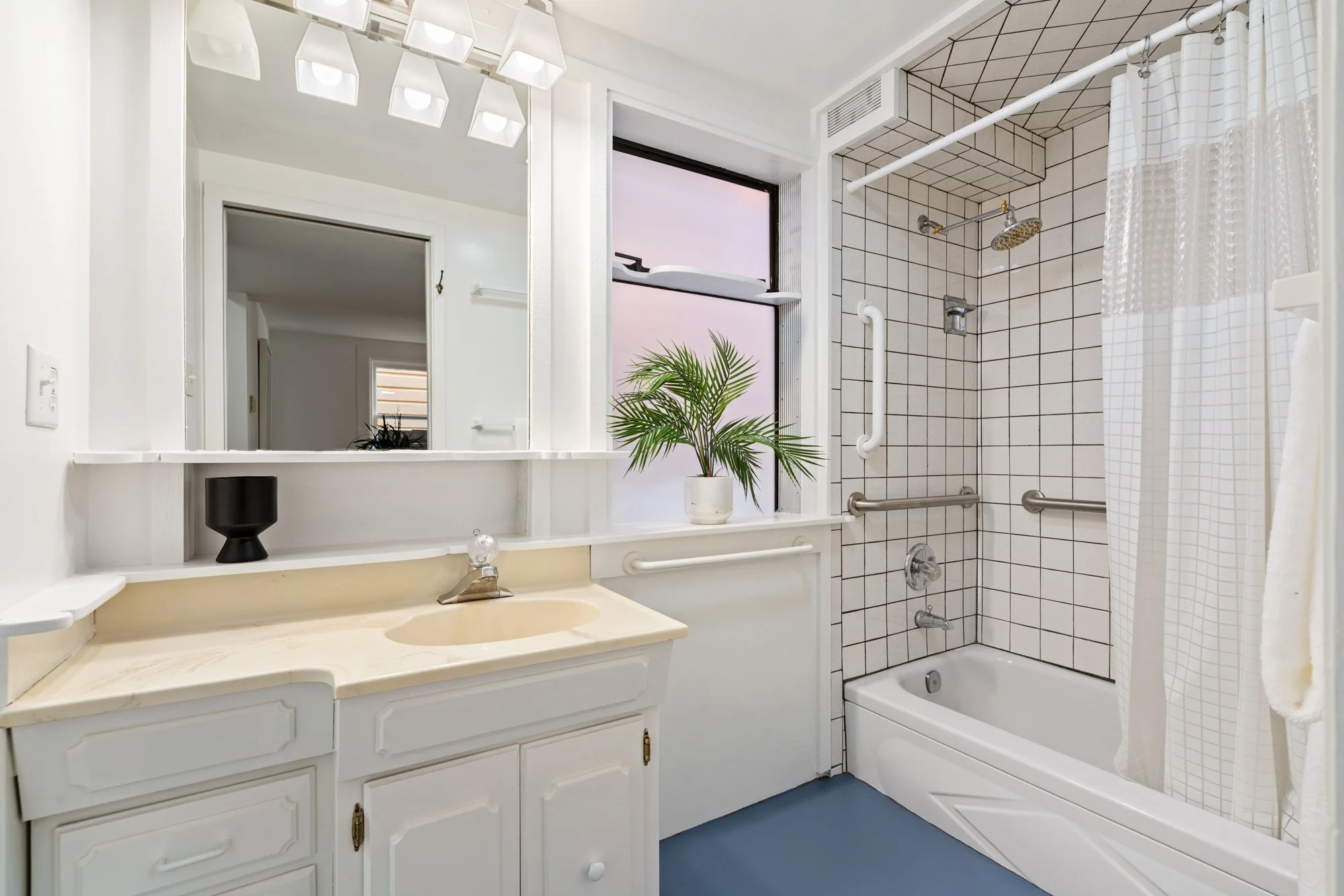 Bathroom with a white vanity, beige countertop, and a large rectangular mirror. There is a black cup on the countertop. A window behind the mirror shows a pinkish sky and a potted plant on the sill. The shower has white tiles with dark grout, a white