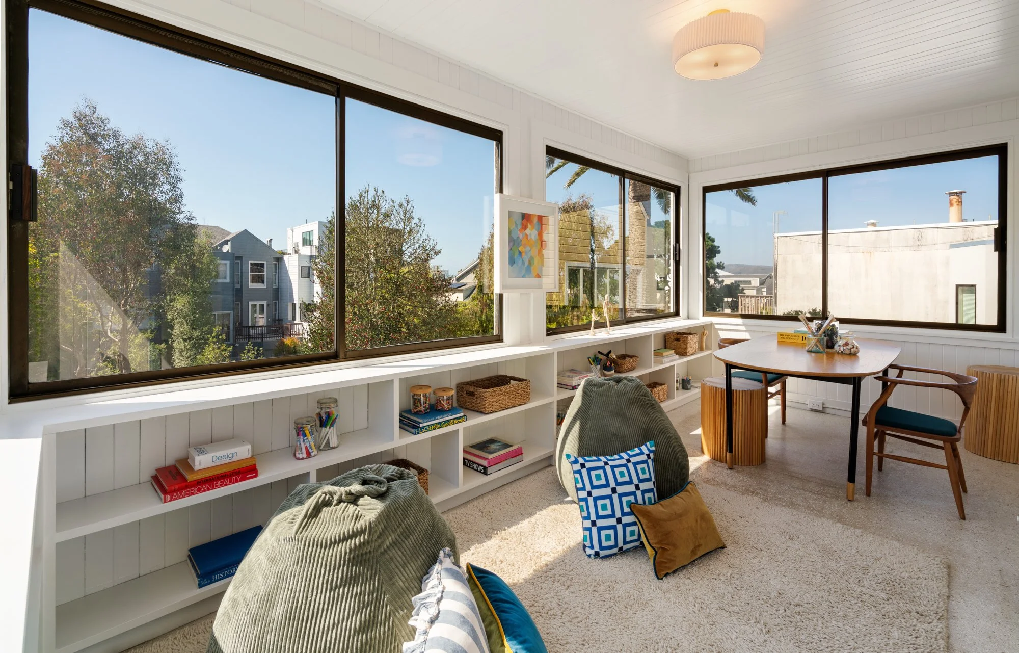 Bright corner room with large windows showing trees and neighboring houses, white bookshelf filled with books and decorative items, armchair with blanket and pillow, small round table with chairs, colorful abstract artwork on wall, and a ceiling ligh