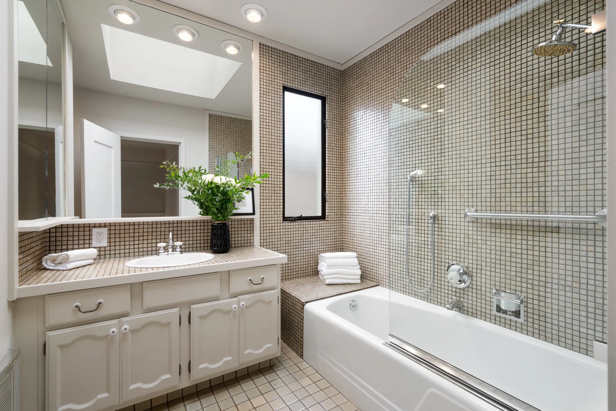 Bathroom with a bathtub, a vanity with a sink, a mirror, and a plant. Tiled walls and floor, window, and skylight.