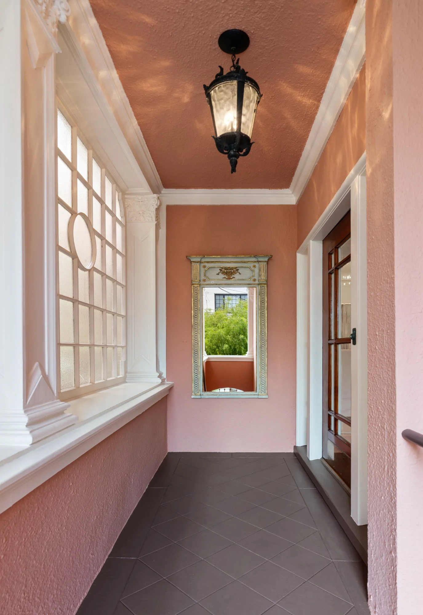 A cozy porch area with pink textured walls, white decorative crown molding, a large window with frosted glass panes, a mirror with a golden ornate frame, and a black hanging lantern light fixture.