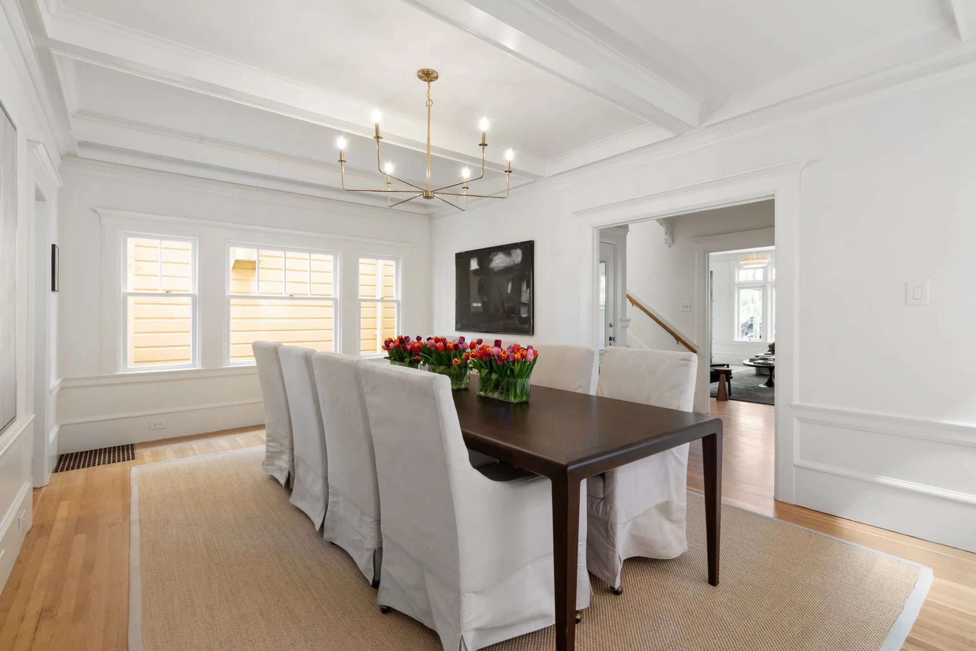 A dining room with a wooden table, white slipcovered chairs, a vase of red and pink tulips, a black wall art behind the table, large windows with white trim, a gold chandelier, light-colored hardwood floors, and a beige area rug.