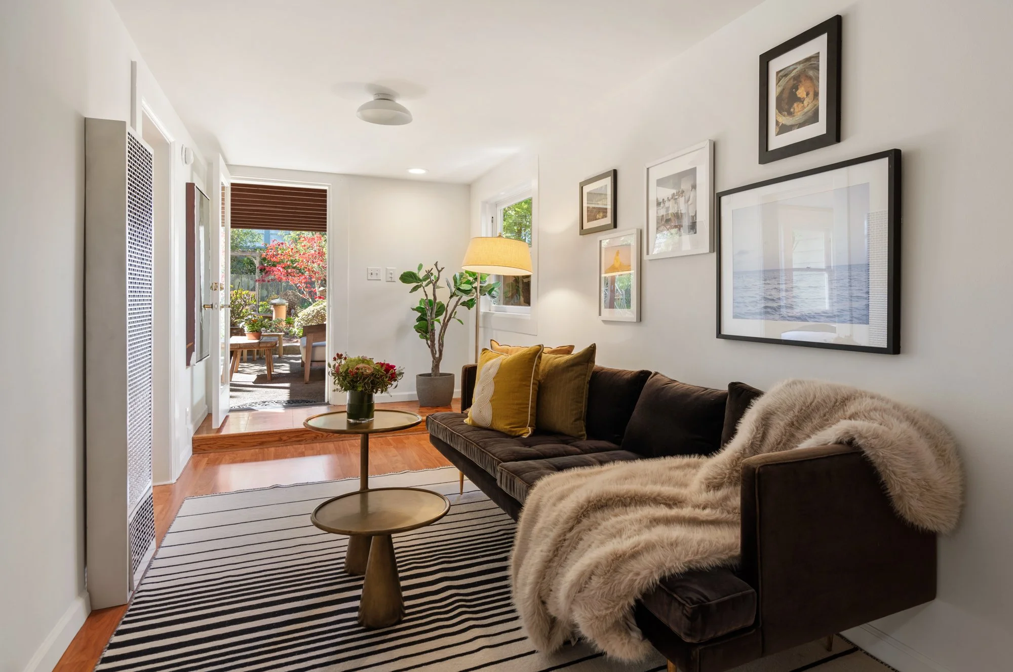 A cozy living room with a black velvet sofa, mustard yellow pillows, and a plush fur throw blanket. There are two small, round wooden coffee tables, a striped rug, a potted plant, a floor lamp, and framed wall art. The room opens to a patio with outd