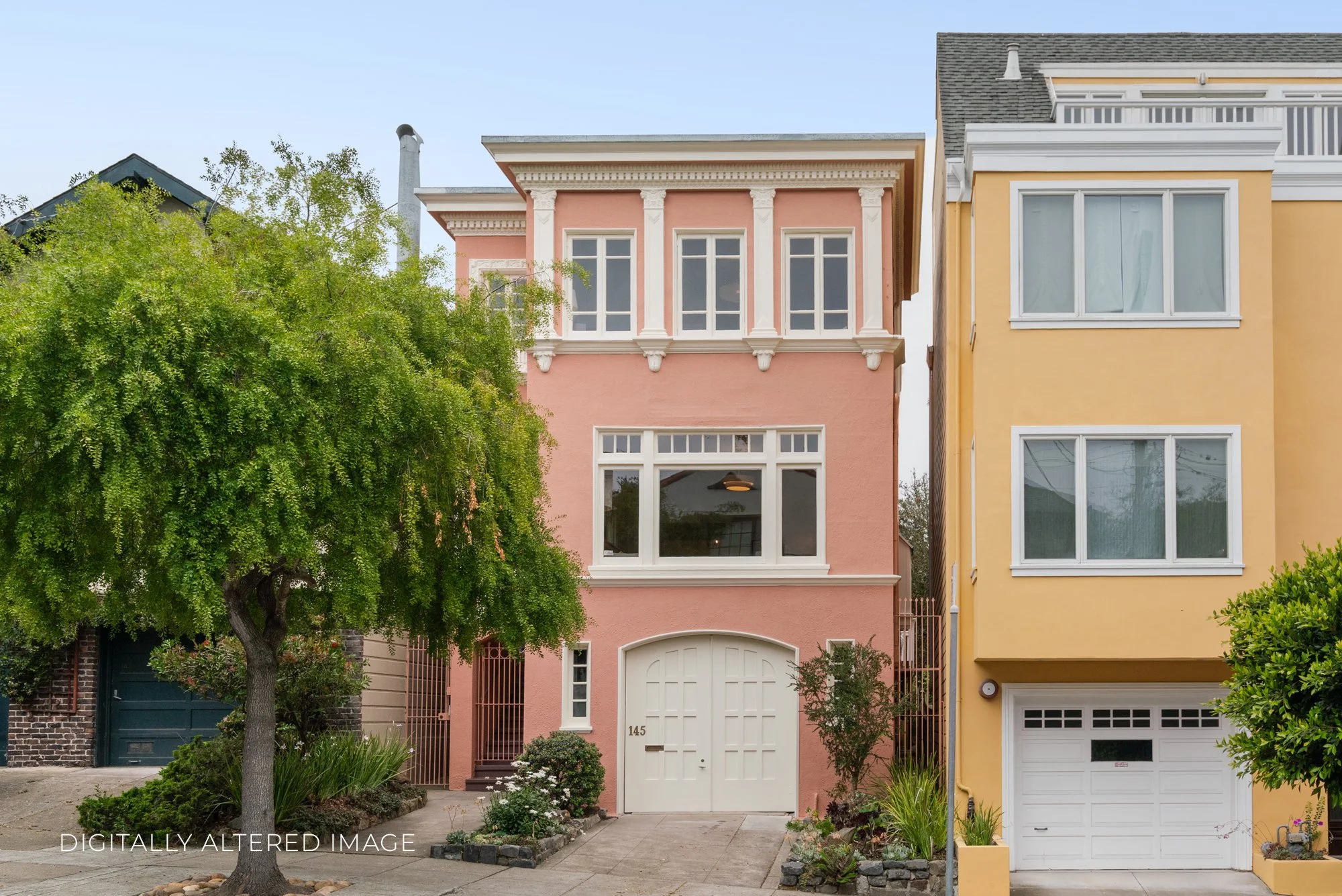 Front view of a colorful pink house with decorative white trim, large front window, white garage door, and lush greenery in front, adjacent to a yellow house with large windows. A large leafy tree partially obscures the pink house. Clear blue sky ove