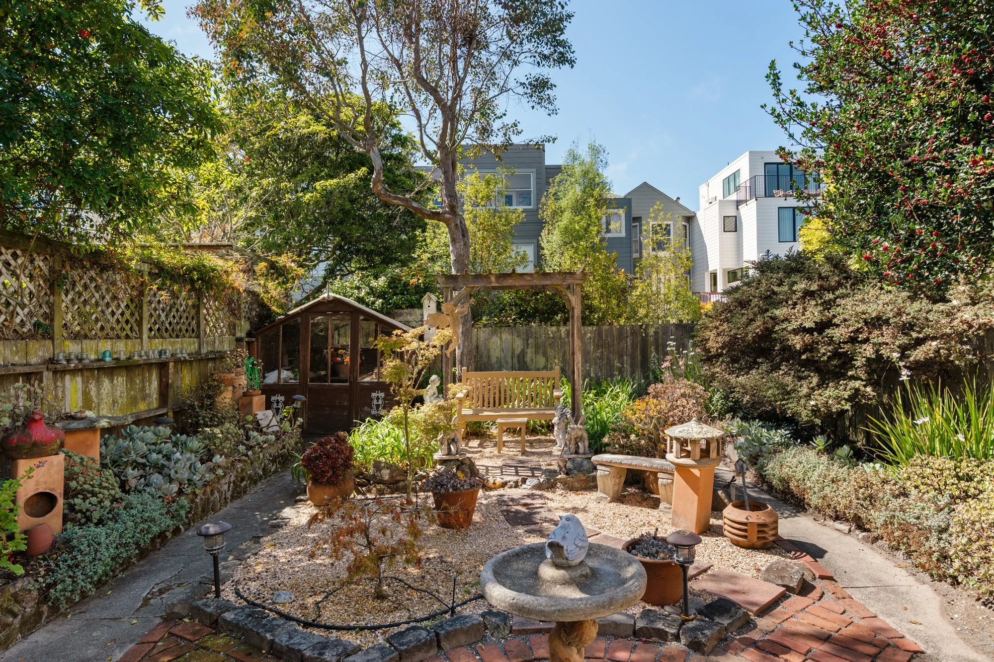 A backyard garden with trees, plants, and decorative items, surrounded by a wooden fence and multi-story residential buildings in the background.