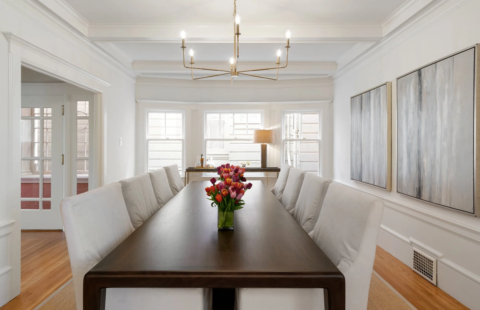 Dining room with a long wooden table, white upholstered chairs, a vase of pink and orange tulips, and modern artwork on the walls. Bright natural light filters through large windows.