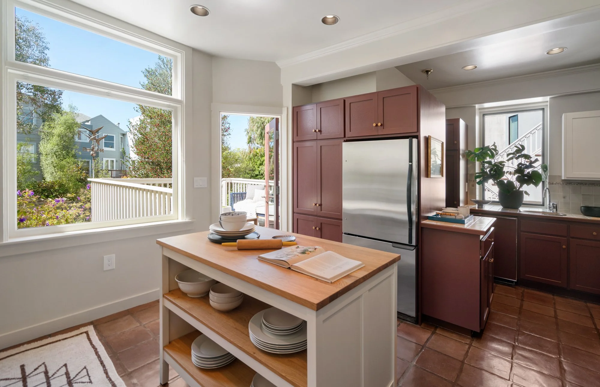 Bright kitchen with large windows overlooking a garden, featuring white and red cabinetry, a stainless steel refrigerator, a wooden island with bowls and dishes, and decorative houseplants, with natural light filling the space.