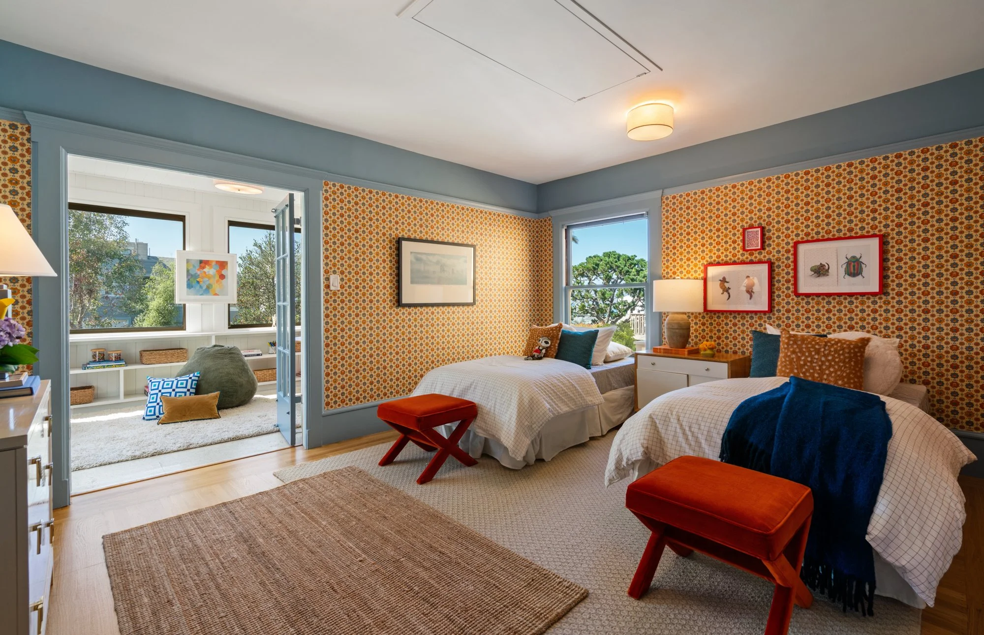 A bedroom with two beds, adorned with pillows and blankets. There are large windows with views of trees outside, and a sunlit porch or sunroom area visible through a doorway. Decor includes framed art on the walls, a table lamp, and a small dresser.