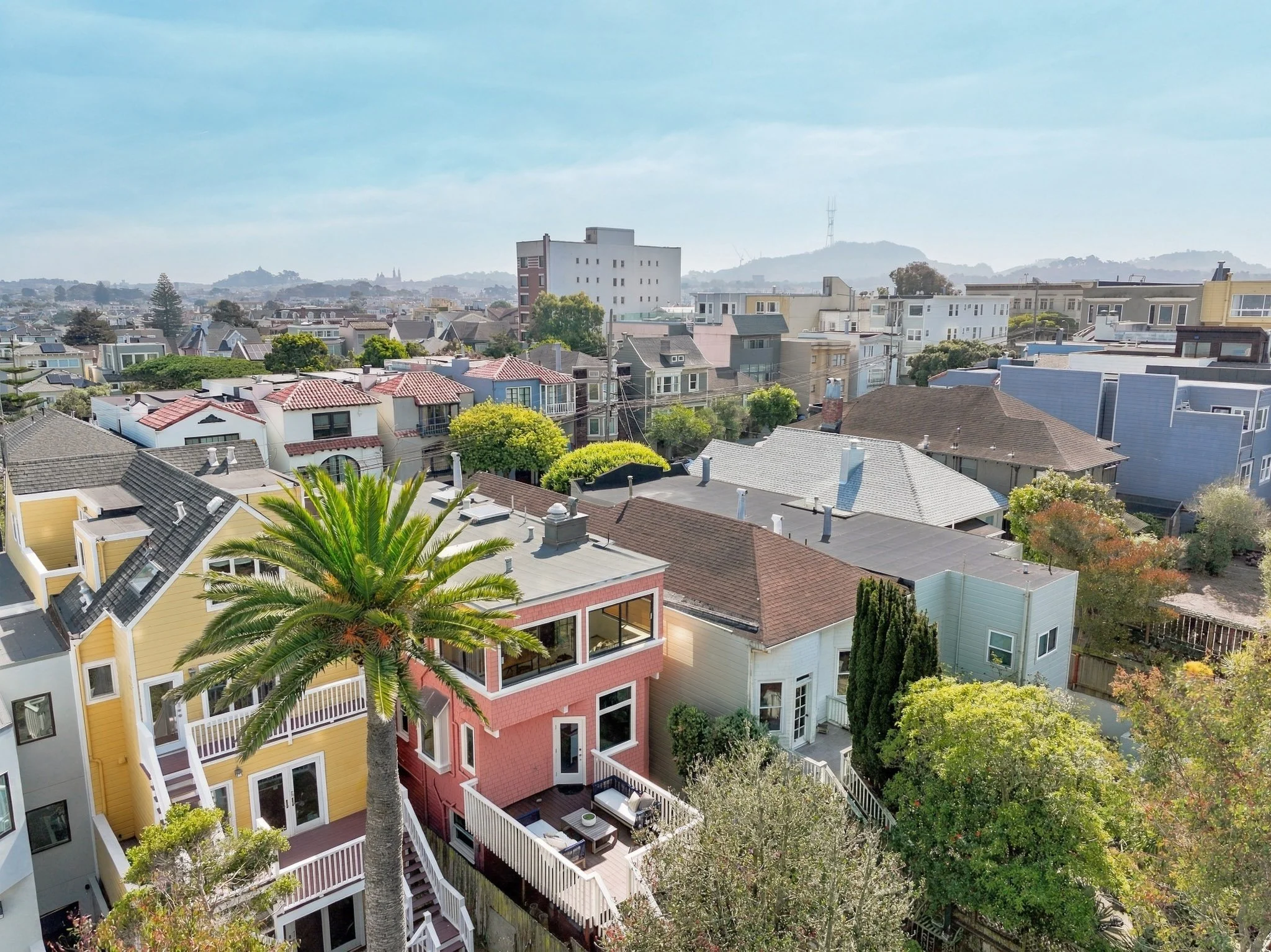 Aerial view of a residential neighborhood with colorful houses, tall trees, and a large hill in the background under a partly cloudy sky.