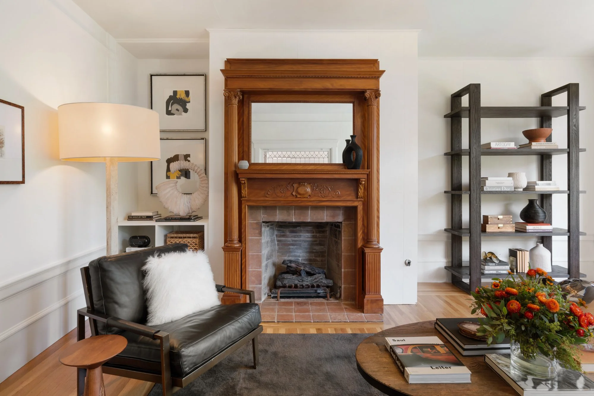 Living room with a fireplace, black leather sofa with a white furry throw, a wooden side table, a large light-colored lamp, and a dark wooden bookshelf displaying books, vases, and decorative objects. There are framed artworks on the wall above the s