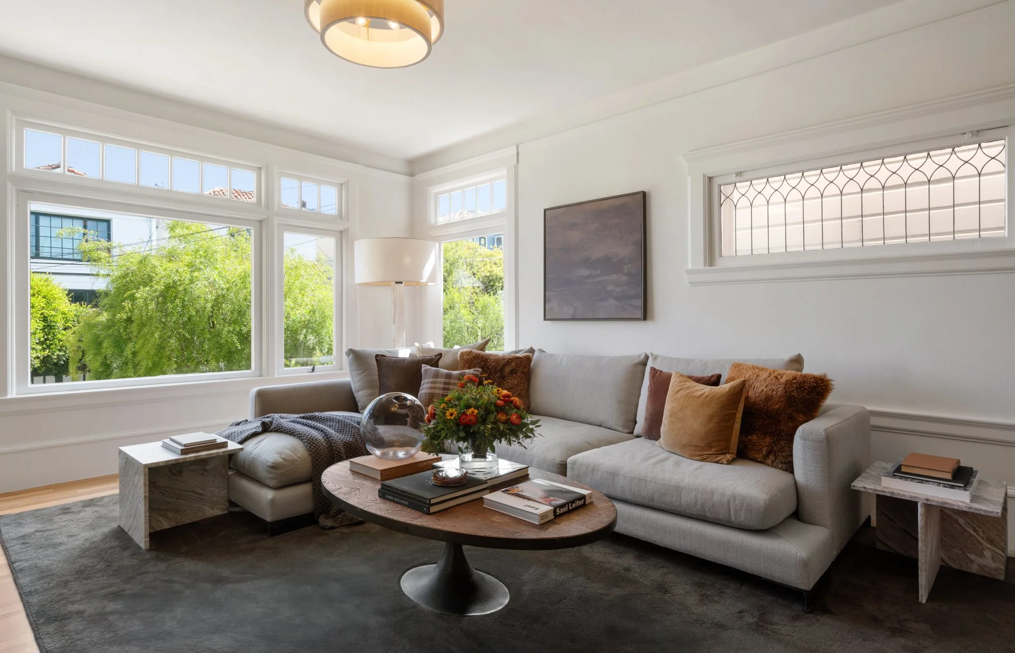 Living room with large windows, white walls, a beige sectional sofa with various pillows, a round coffee table with books and a flower arrangement, a marble side table, and a floor lamp. There is a ceiling light fixture and outside greenery visible t