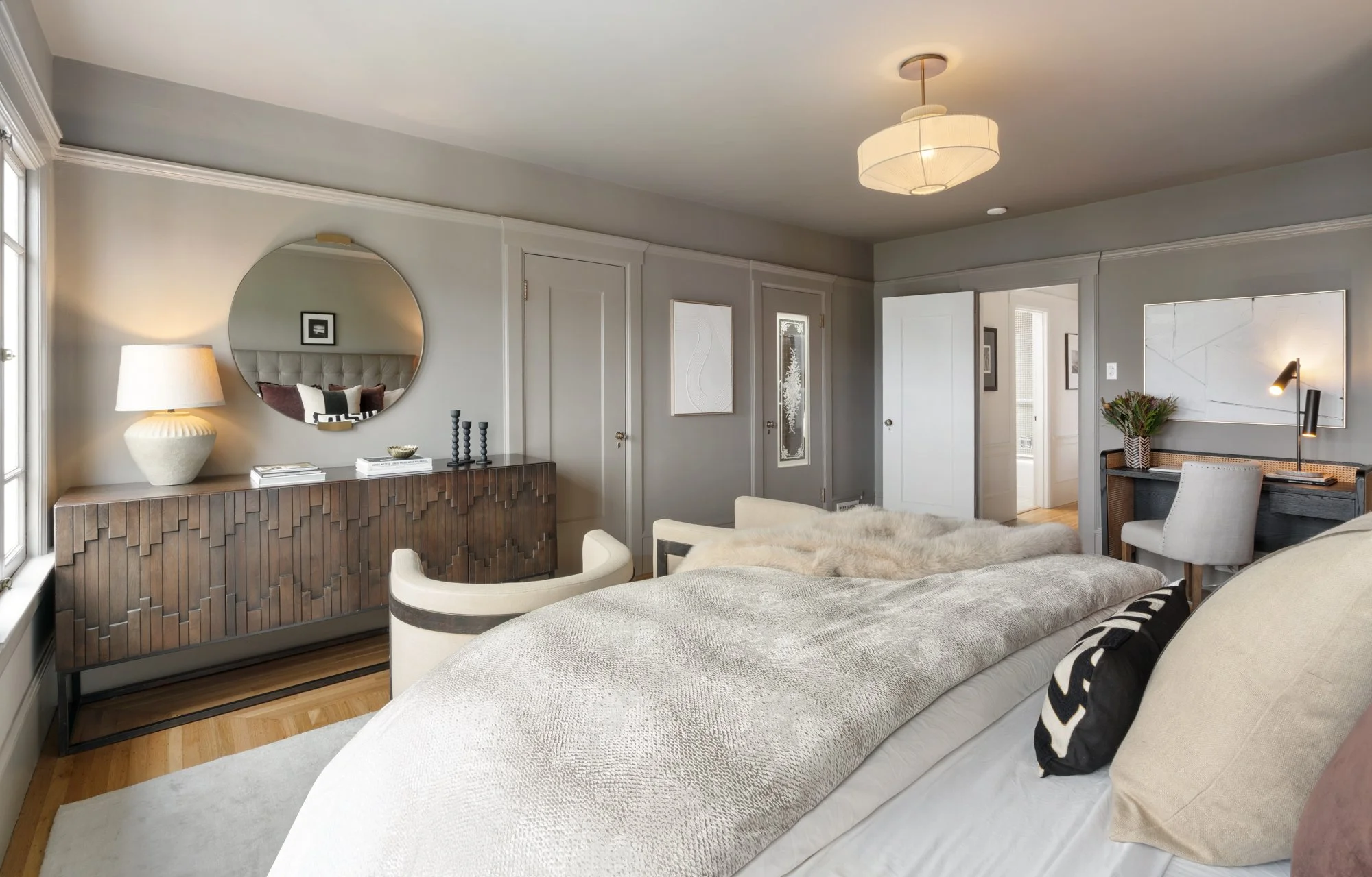A cozy bedroom with a bed, armchair, and side table. A large window brings in natural light, and a decorative cabinet with a lamp and artwork is on the left side. The room features neutral colors and modern decor.