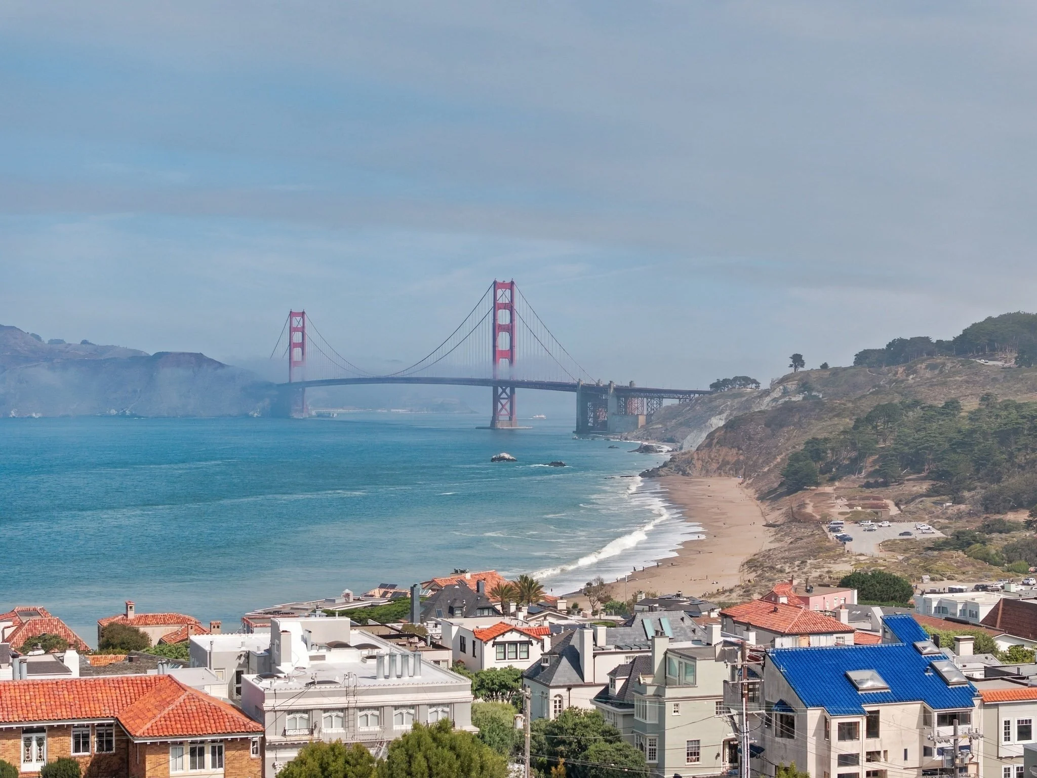 A view of the Golden Gate Bridge in San Francisco, California, with houses and buildings along the coastline, and a beach with parked cars in the foreground.