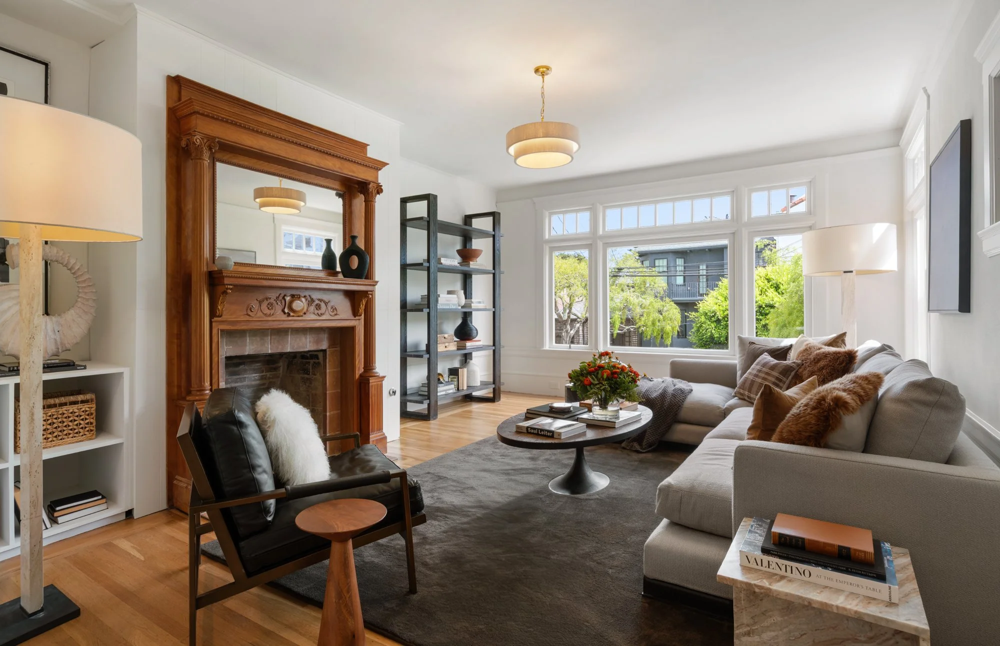Bright living room with large windows, a white sofa adorned with fur pillows, a black armchair with a fur throw, a wooden coffee table with books and a floral arrangement, a fireplace with a wooden mantle, and black shelving with decorative items. Modern lighting fixtures are hanging from the ceiling.