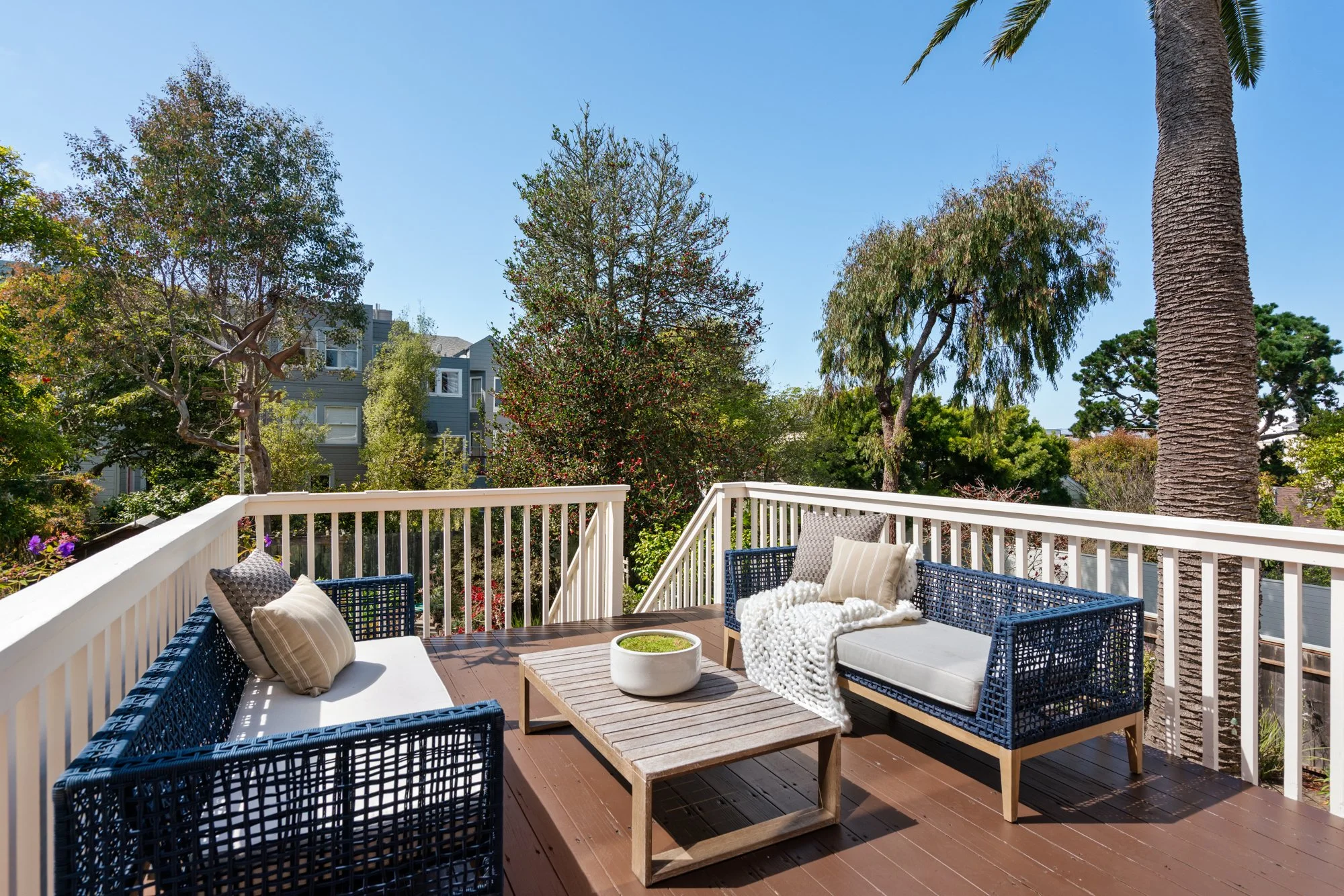 A balcony with outdoor seating, including blue and white woven sofas with pillows, a wooden table with a decorative bowl, overlooking lush green trees and a blue sky.