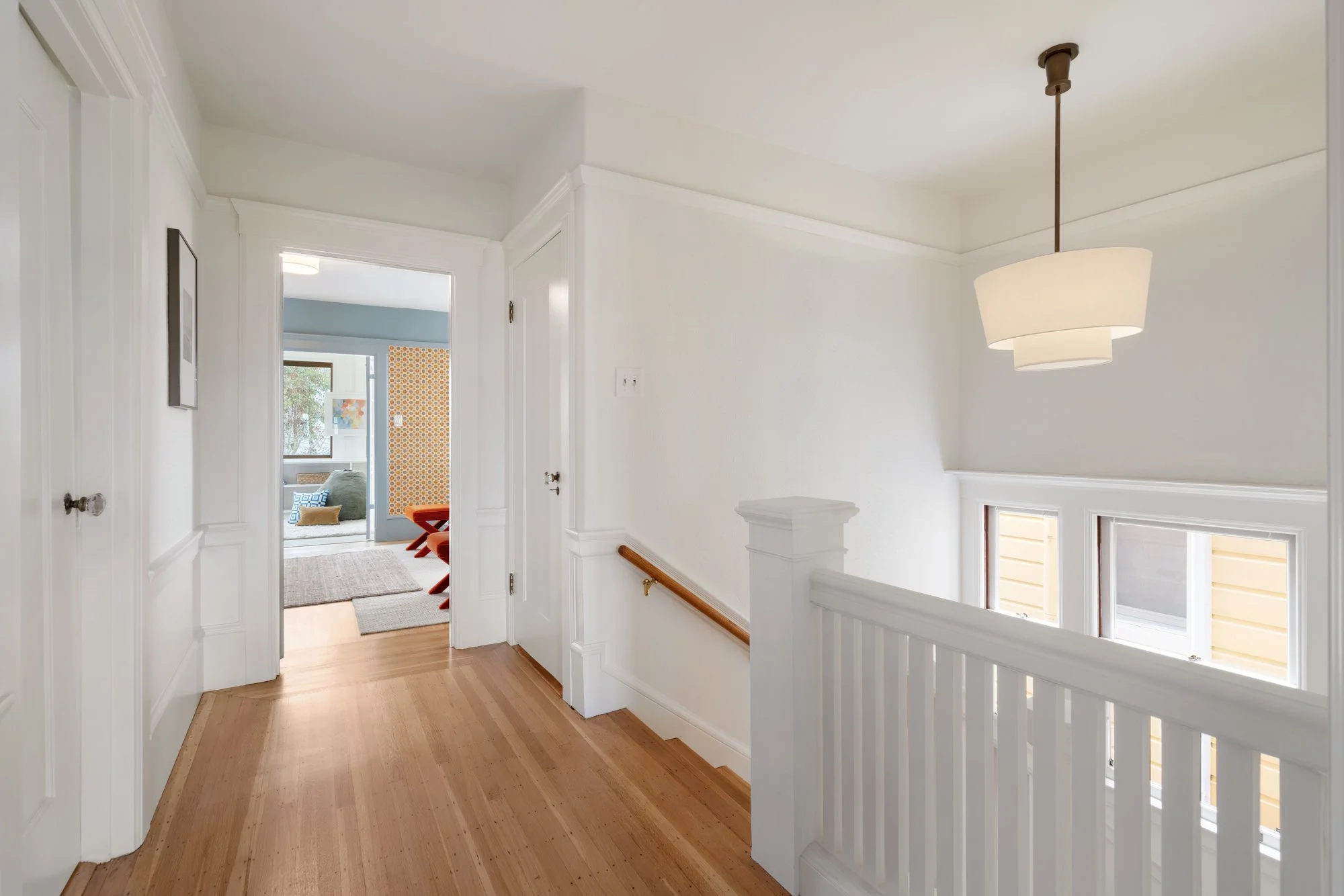 A hallway with white walls, hardwood flooring, a curved railing, and a hanging ceiling lamp. A doorway leads to a living room with blue walls and colorful furniture.