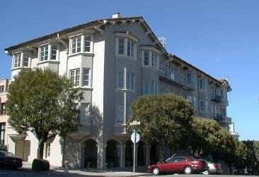 A multi-story residential building with rounded windows and balconies, located on a street corner with trees and parked cars.