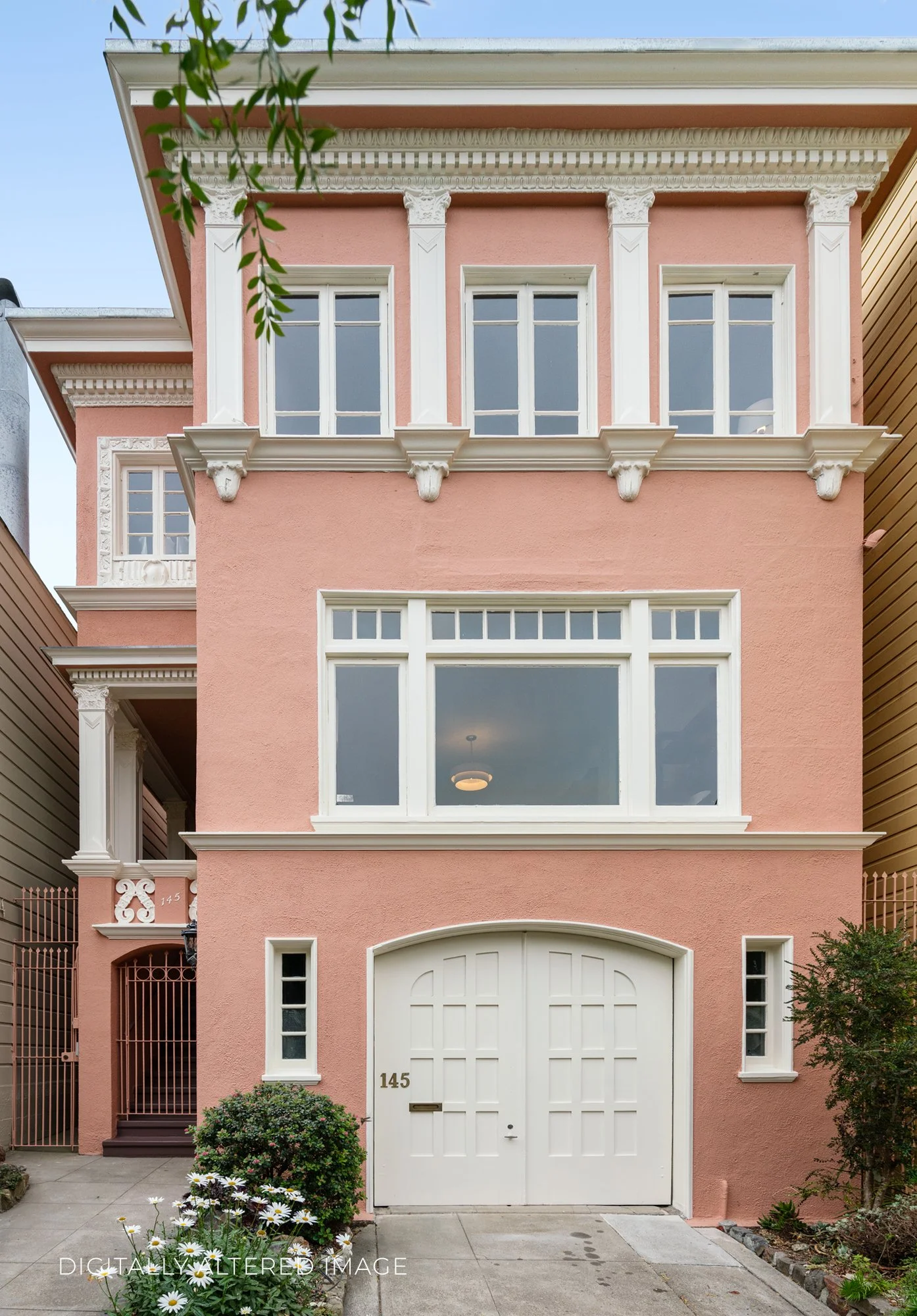 A pink three-story house with white decorative trim, upper-story windows, and a white arched front door. The house number 145 is displayed on the door. There is greenery and flowers in front, with a small bush and daisies. The house is enclosed by a 