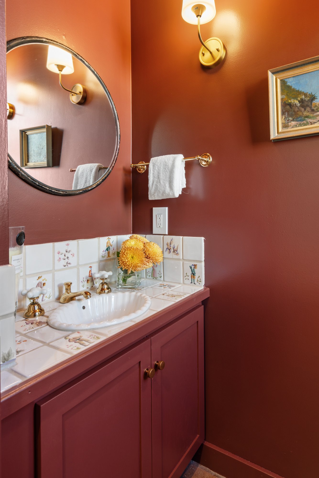 A bathroom vanity with a white sink, vintage gold fixtures, and a tiled countertop adorned with decorative tiles and a bouquet of yellow flowers. There is a round mirror on a dark red wall, complemented by a wall sconce light fixture and a framed pai