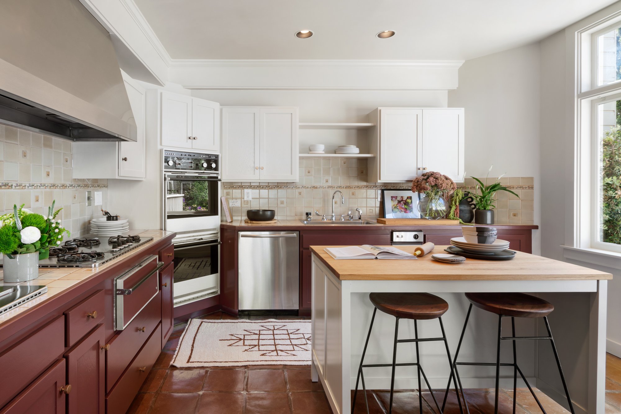 A bright kitchen with white cabinets, a tiled beige backsplash, a stove, a sink, various plants and flowers on the counter, open shelving with plates, a window bringing in natural light, a kitchen island with two stools, and a decorative rug on the terracotta tile floor.