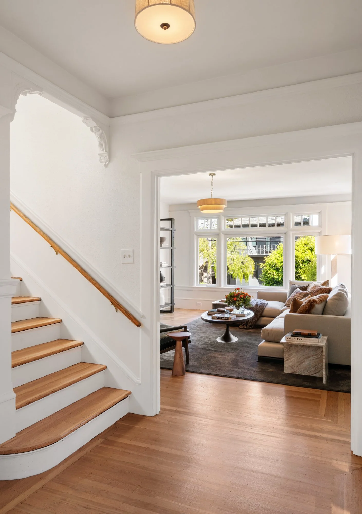 Living room with large window, beige sofa with throw pillows, coffee table with flowers and books, black bookshelf, and a modern ceiling light, view of a balcony with greenery outside.