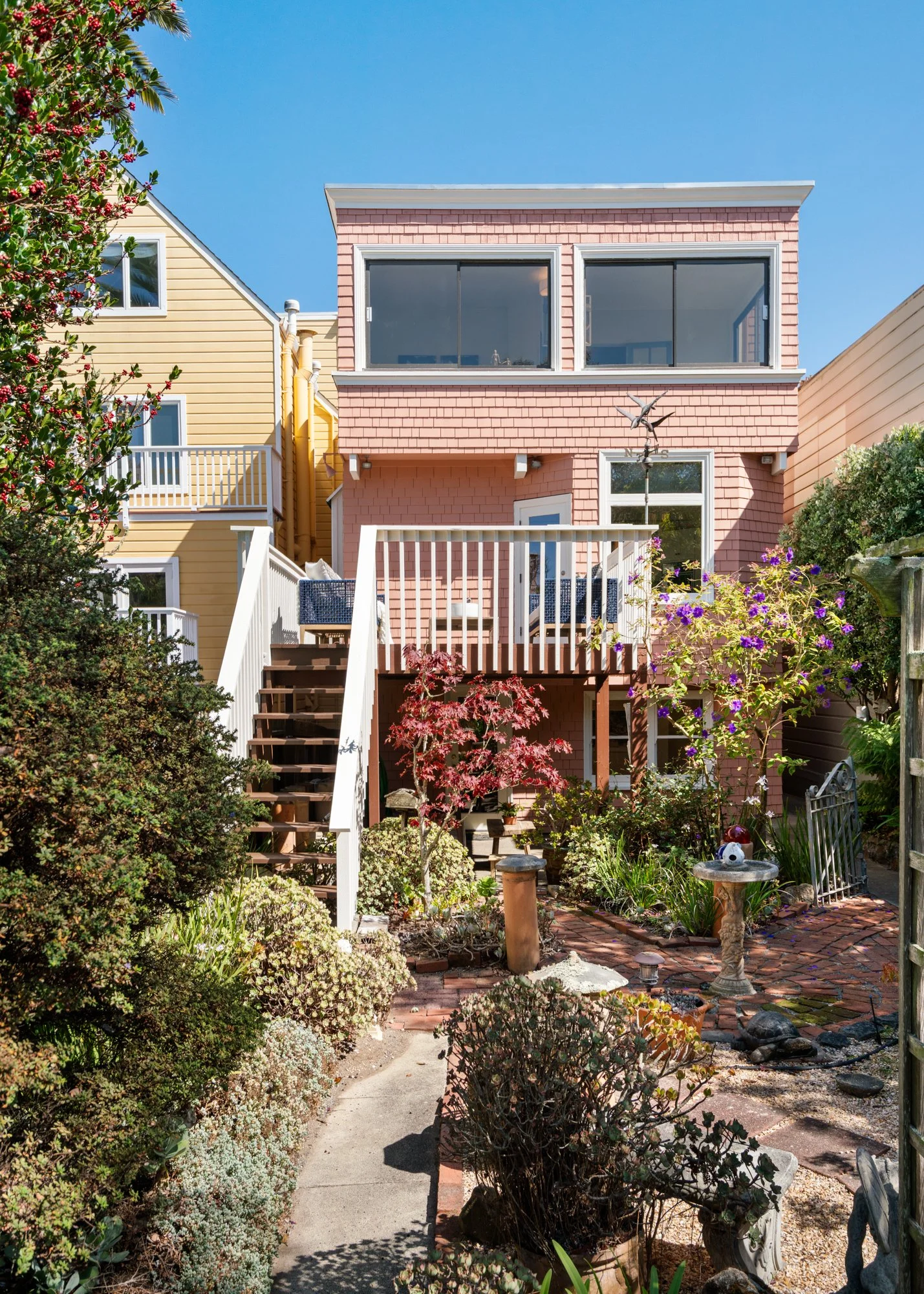 Colorful backyard garden with plants, shrubs, and trees, brick pathway, decorative birdbath, and a staircase leading to a pink two-story house with a balcony, under a clear blue sky.