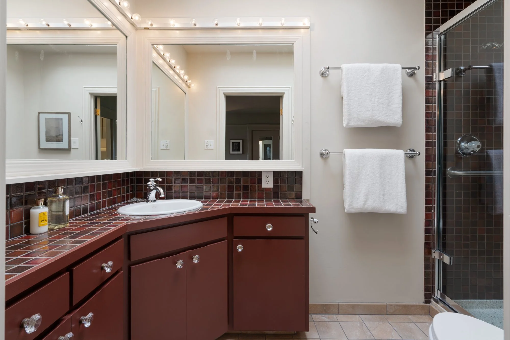 Bathroom with vanity and large mirror, white towels on a towel rack, tiled shower, and framed pictures on the wall.