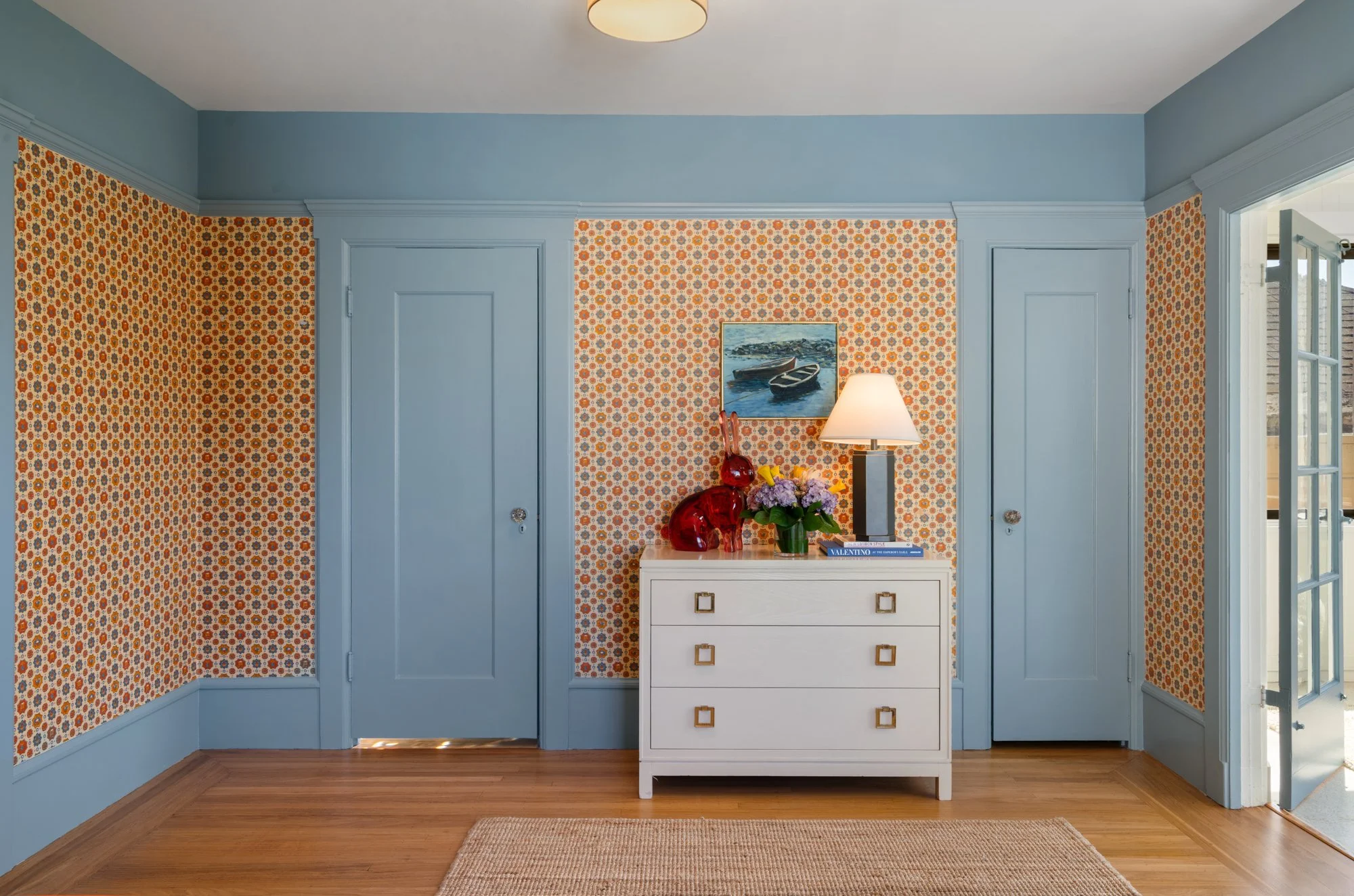 A hallway with light blue walls and patterned wallpaper on the back wall. There is a white dresser with gold square handles, topped with decorative red glass vases, a vase of purple flowers, and a table lamp. Above the dresser, a framed painting of b