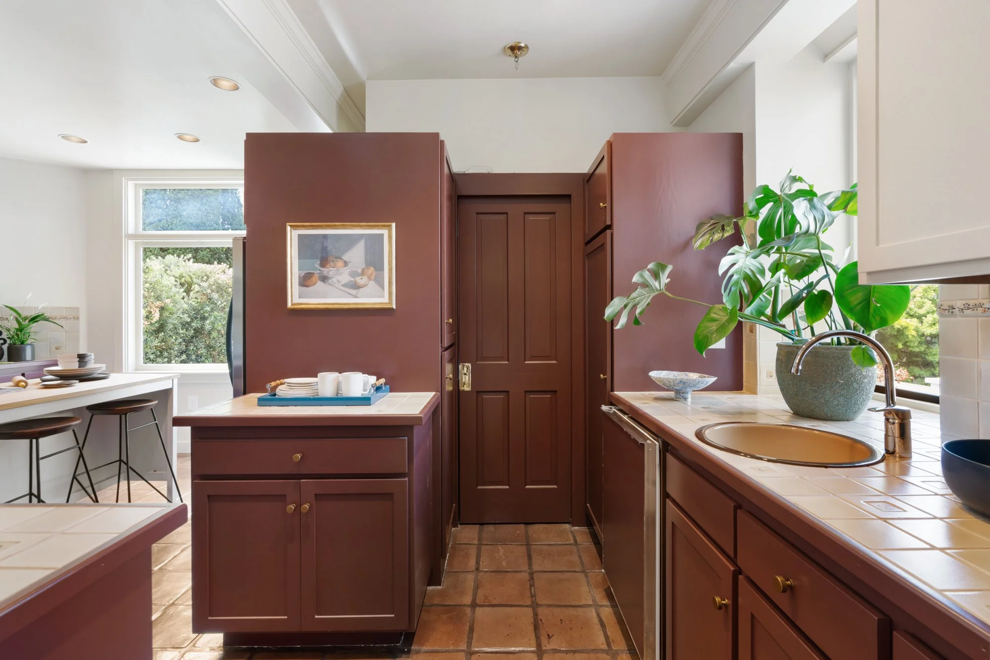 A cozy kitchen with a large window showing greenery outside, featuring brown cabinets, a tiled countertop, a large potted monstera plant, and a framed picture on a partition wall.