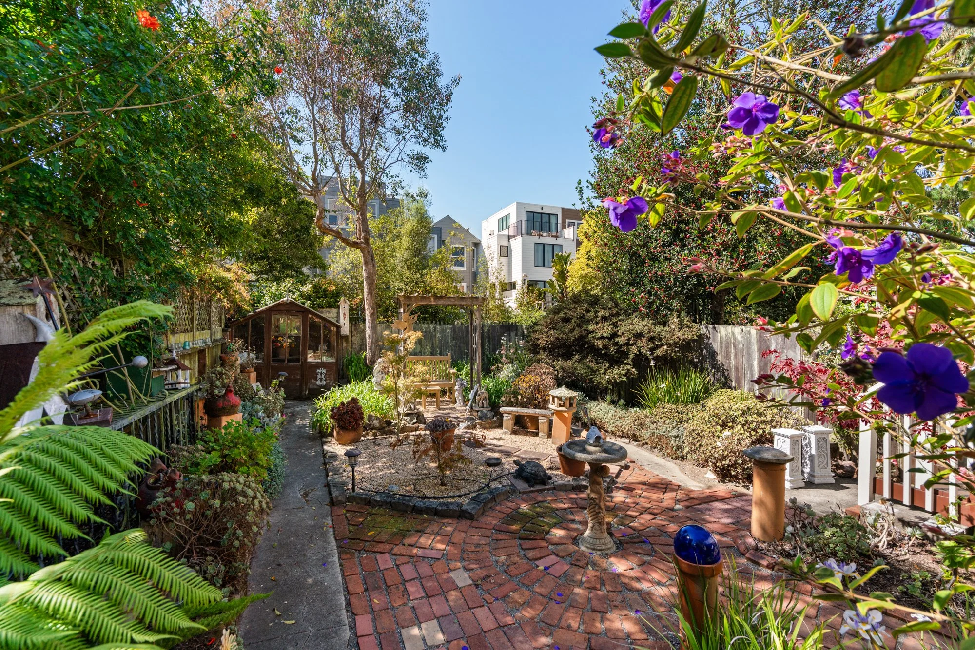 A lush backyard garden with a brick pathway, surrounded by potted plants, small statues, and a greenhouse, with trees and flowering bushes in the background under a clear blue sky.