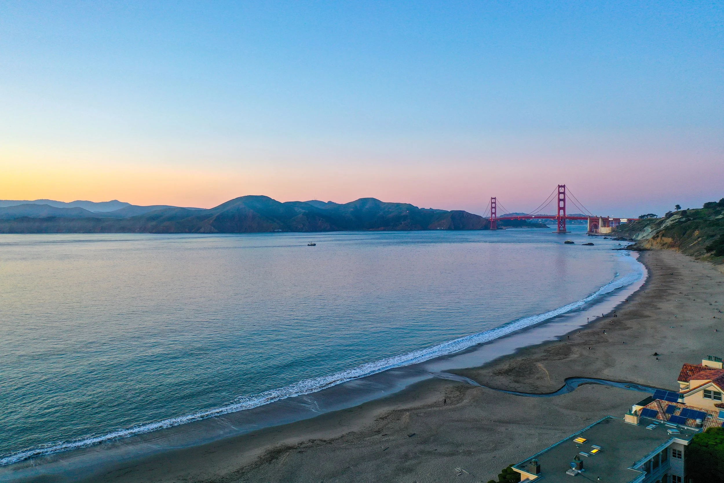 A view of the Golden Gate Bridge in San Francisco at sunset, with calm water, distant hills, and some houses near the shoreline.