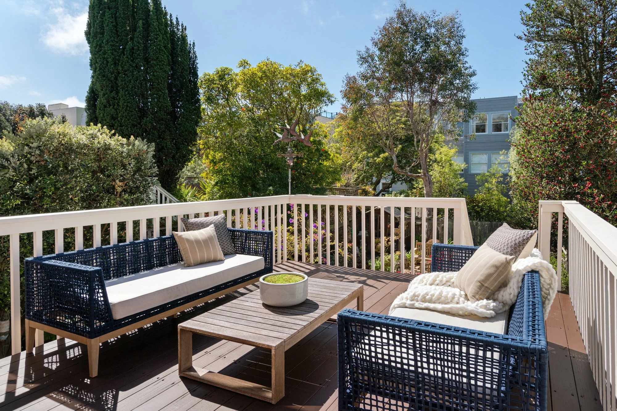 Outdoor patio with blue wicker sofas and beige pillows, a wooden coffee table with a white planter, surrounded by greenery and trees under a clear blue sky.