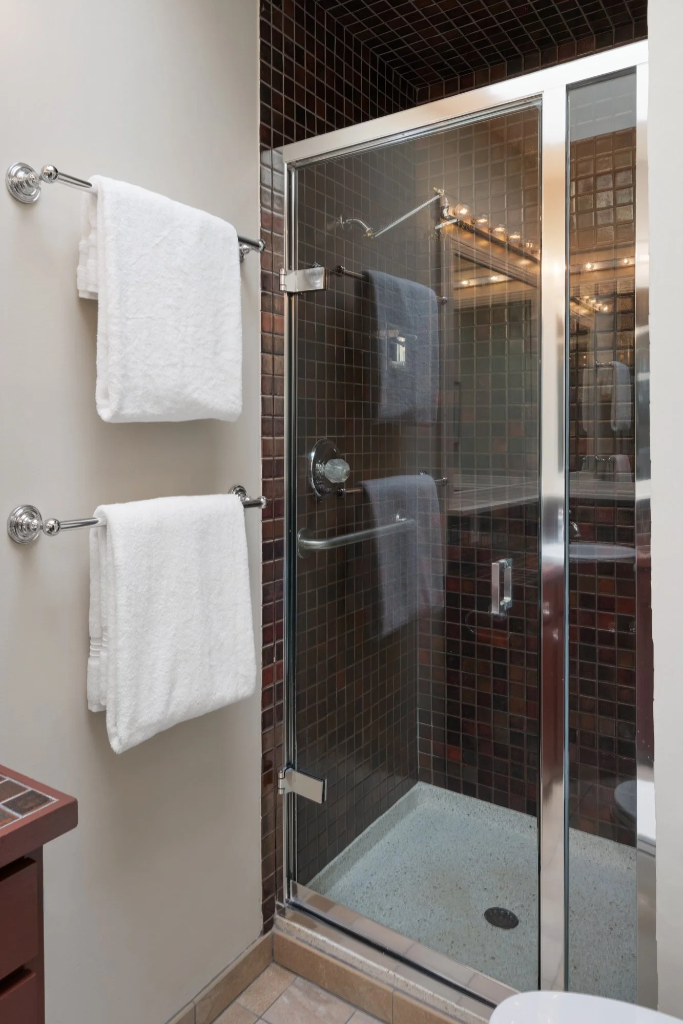 Small shower with dark tiles, glass door, and towel racks with white towels.