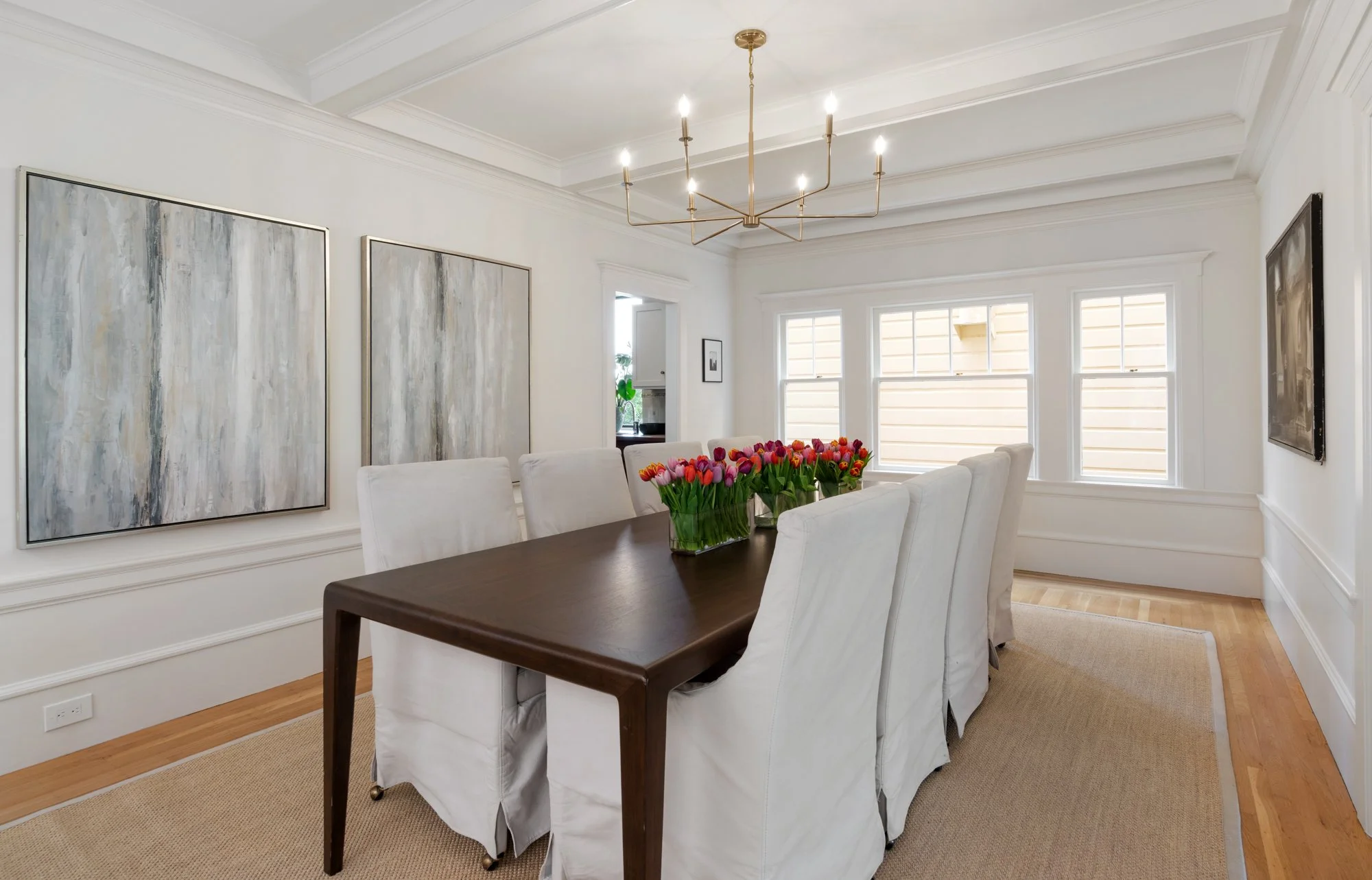 Dining room with white chairs around a dark wooden table, a vase of pink and red tulips, large abstract paintings on white walls, three windows, and a modern chandelier.