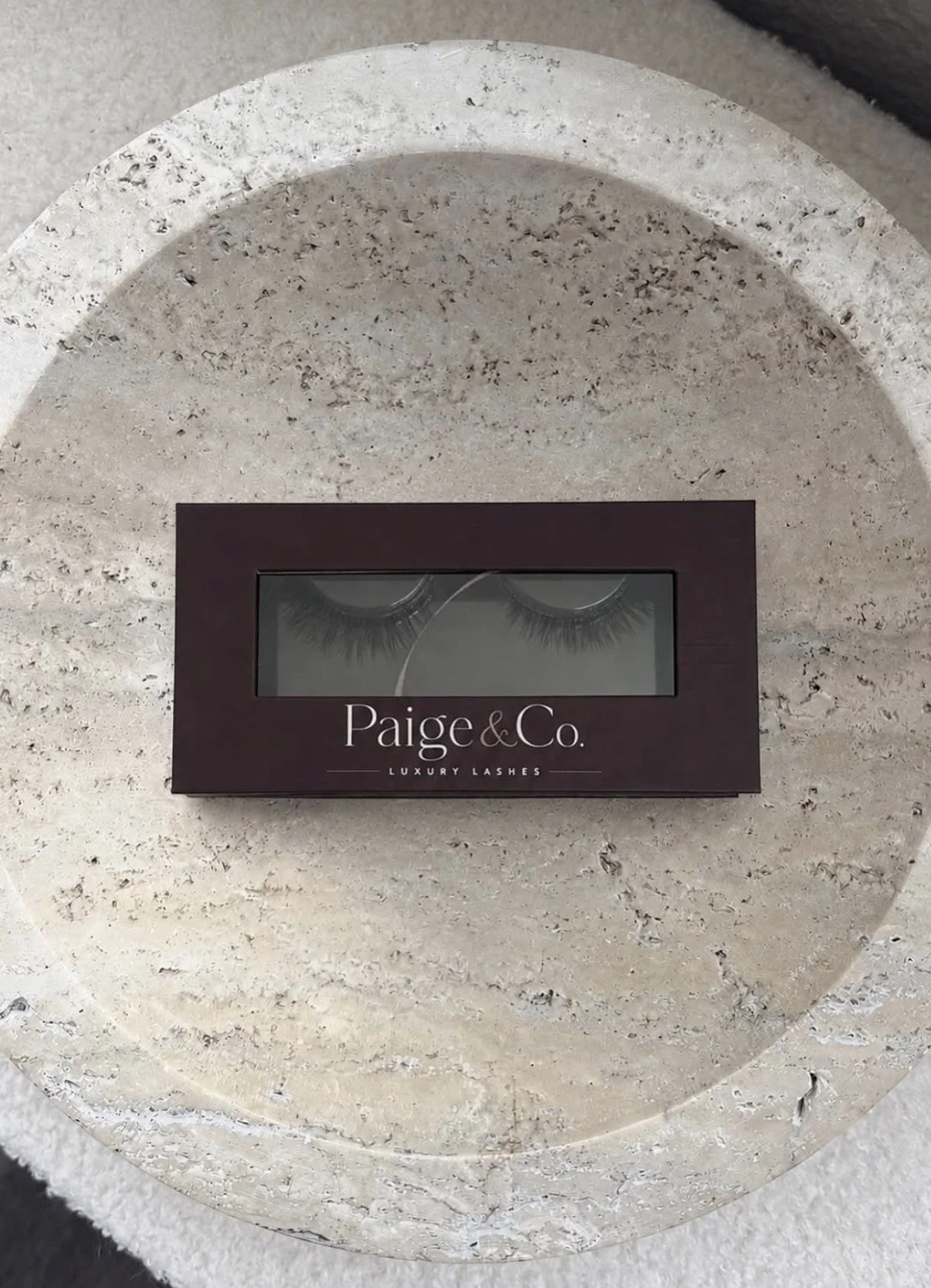 Box of luxury false eyelashes labeled "Paige & Co." resting on a textured stone circular surface.