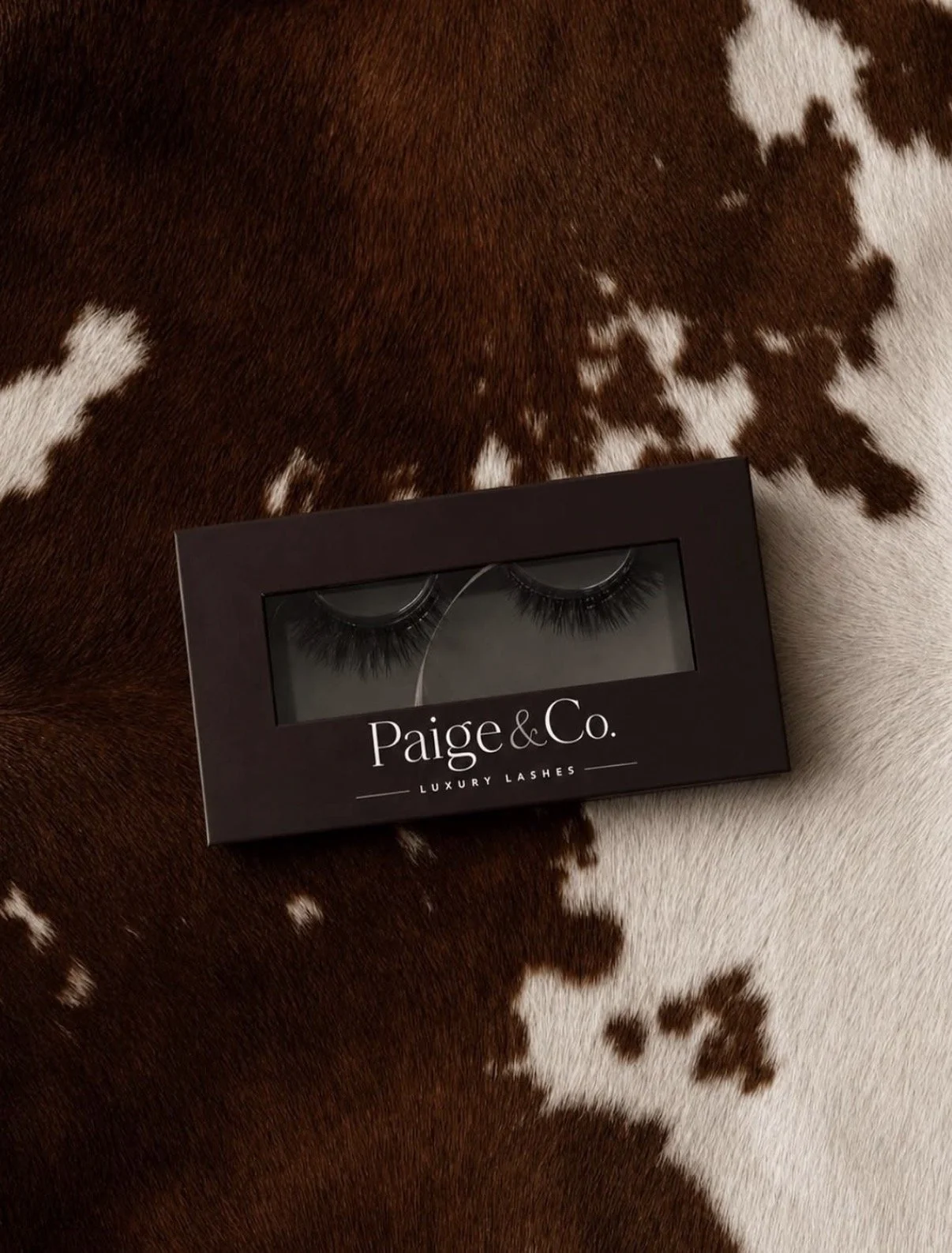 A box of Paige & Co. luxury false eyelashes on a cowhide surface.