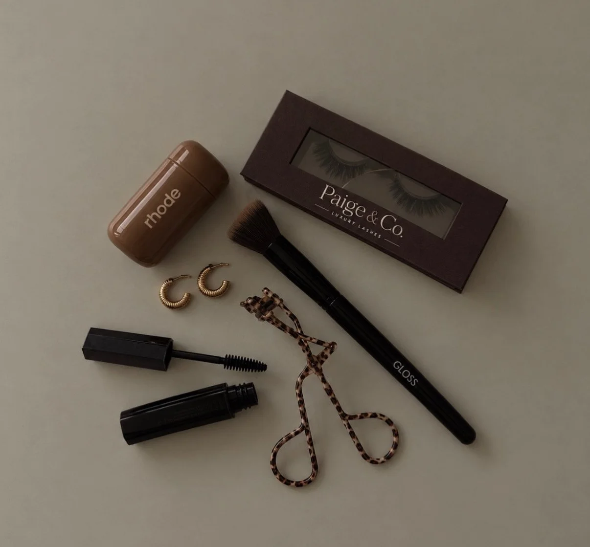Makeup and beauty accessories including a box of false eyelashes, a makeup brush, a tube of mascara, a small container of Rhode skincare product, a pair of gold hoop earrings, a tortoise shell hair clip, a pair of eyelash curlers, and a black lip gloss.